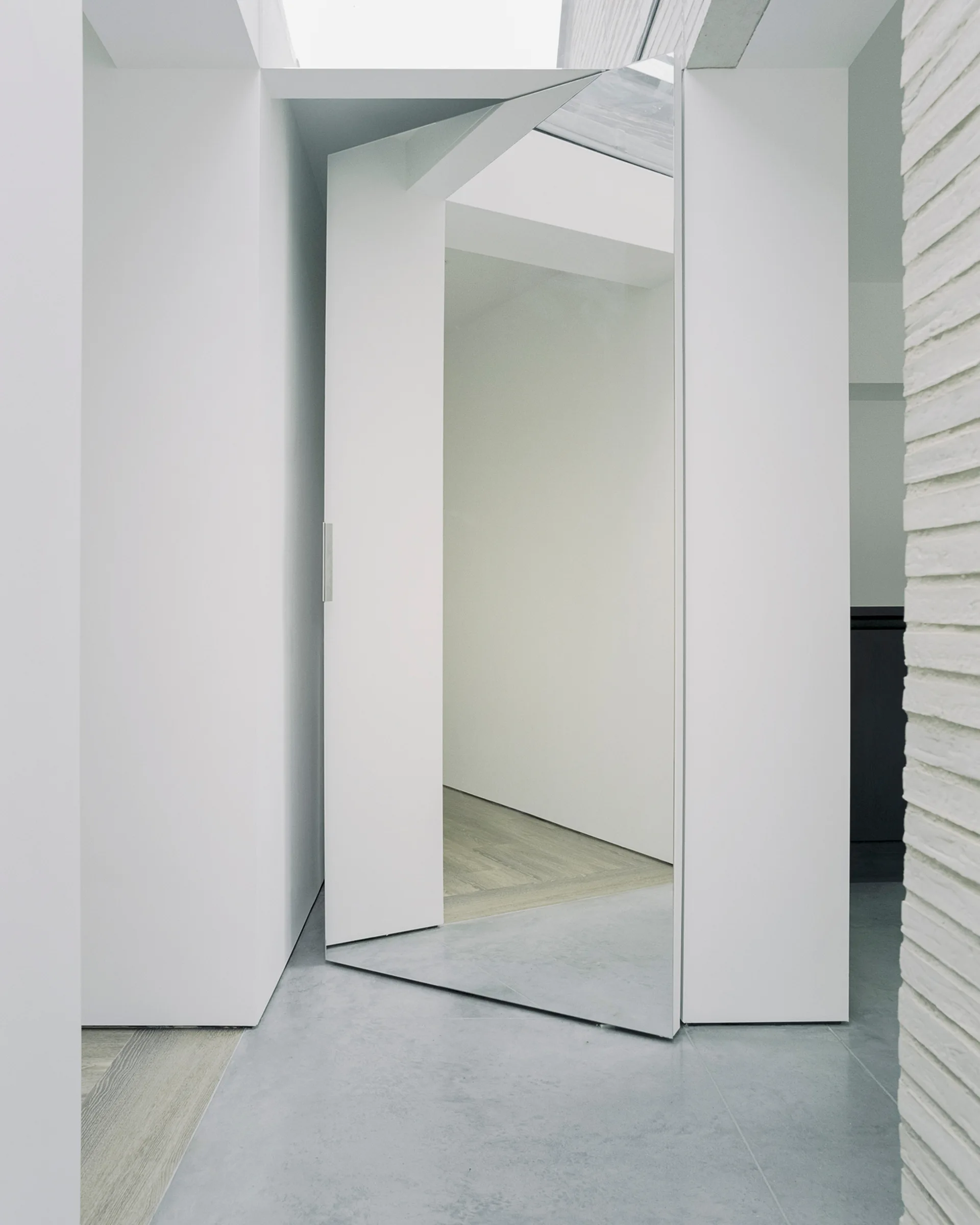 A full-height pivoting mirrored door stands open between matte white walls and a painted brick wall in a minimalist interior.