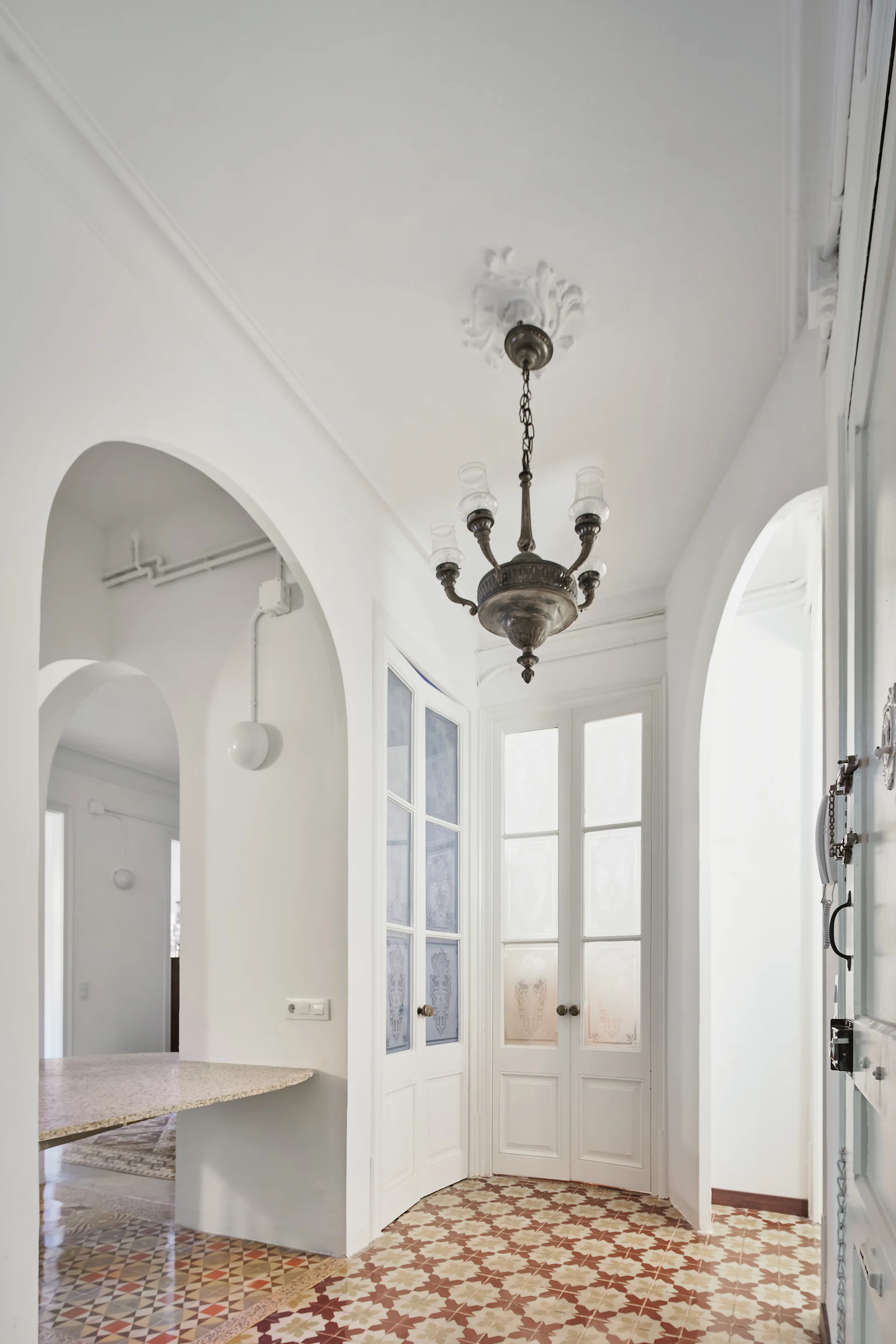 A white hallway features classic arched doorways, intricate patterned floor tiles in warm tones, and a vintage metal chandelier.
