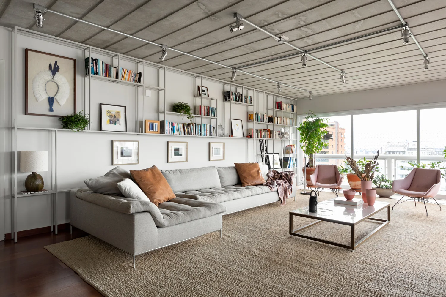 An industrial modern living room features a gray sectional sofa, a large metal wall shelving unit, and a ribbed concrete ceiling with track lighting.