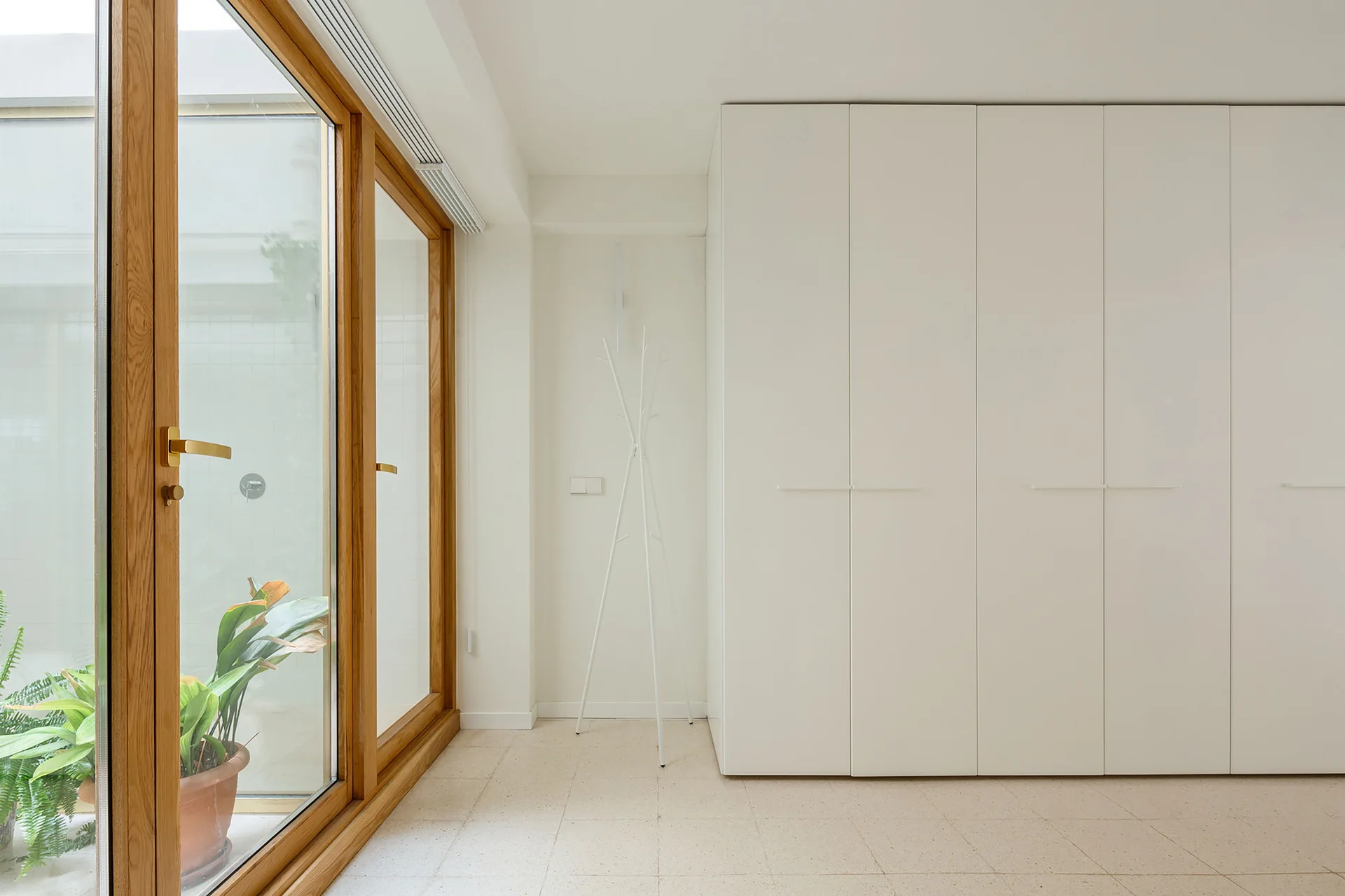 A minimalist hallway features white floor-to-ceiling cabinets with integrated handles, a slim white coat rack, and large wood-framed glass doors.