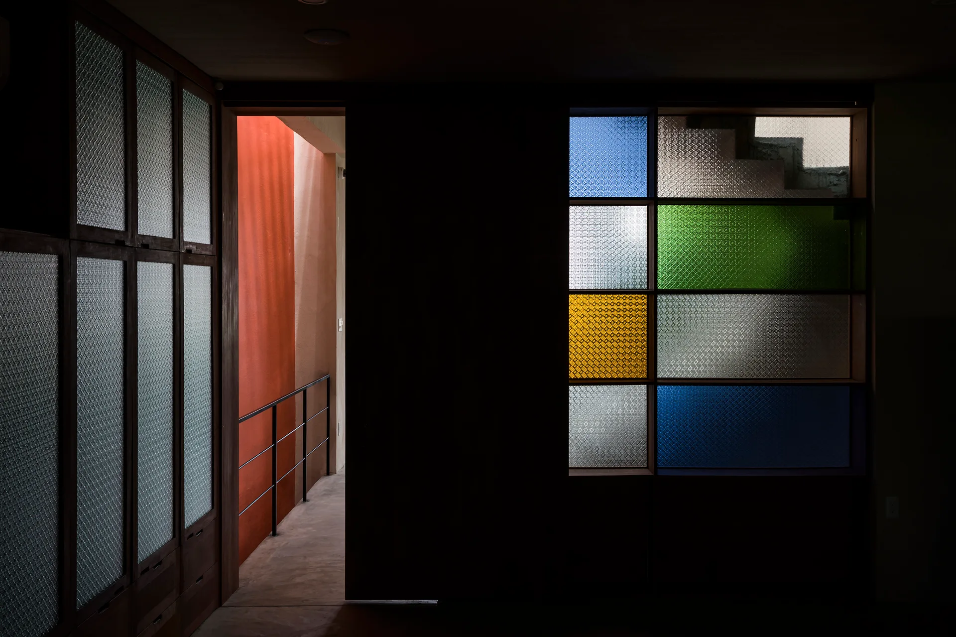 A dark interior featuring a wooden built-in wardrobe, an open doorway showing a bright orange corridor, and a partition with colored textured glass.