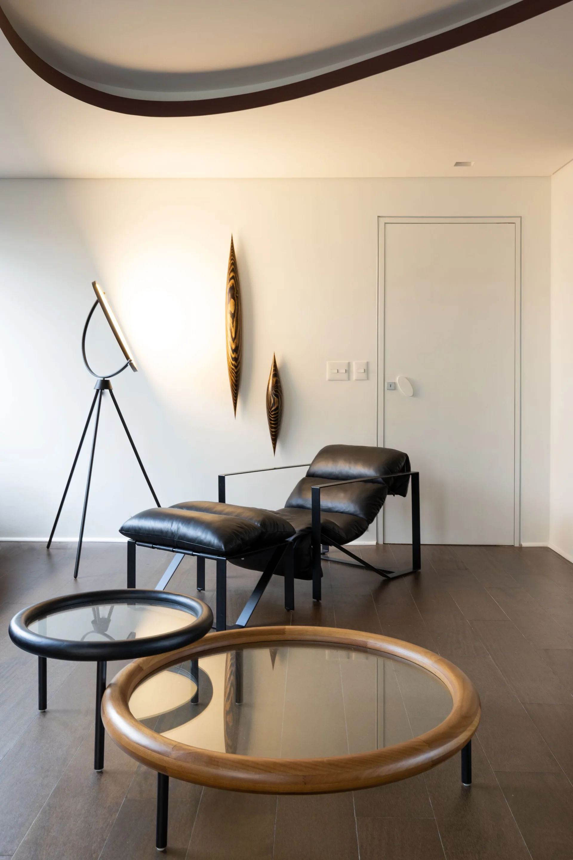 A minimalist living room corner features a black leather lounge chair, a metal tripod floor lamp, and two round glass-topped coffee tables.