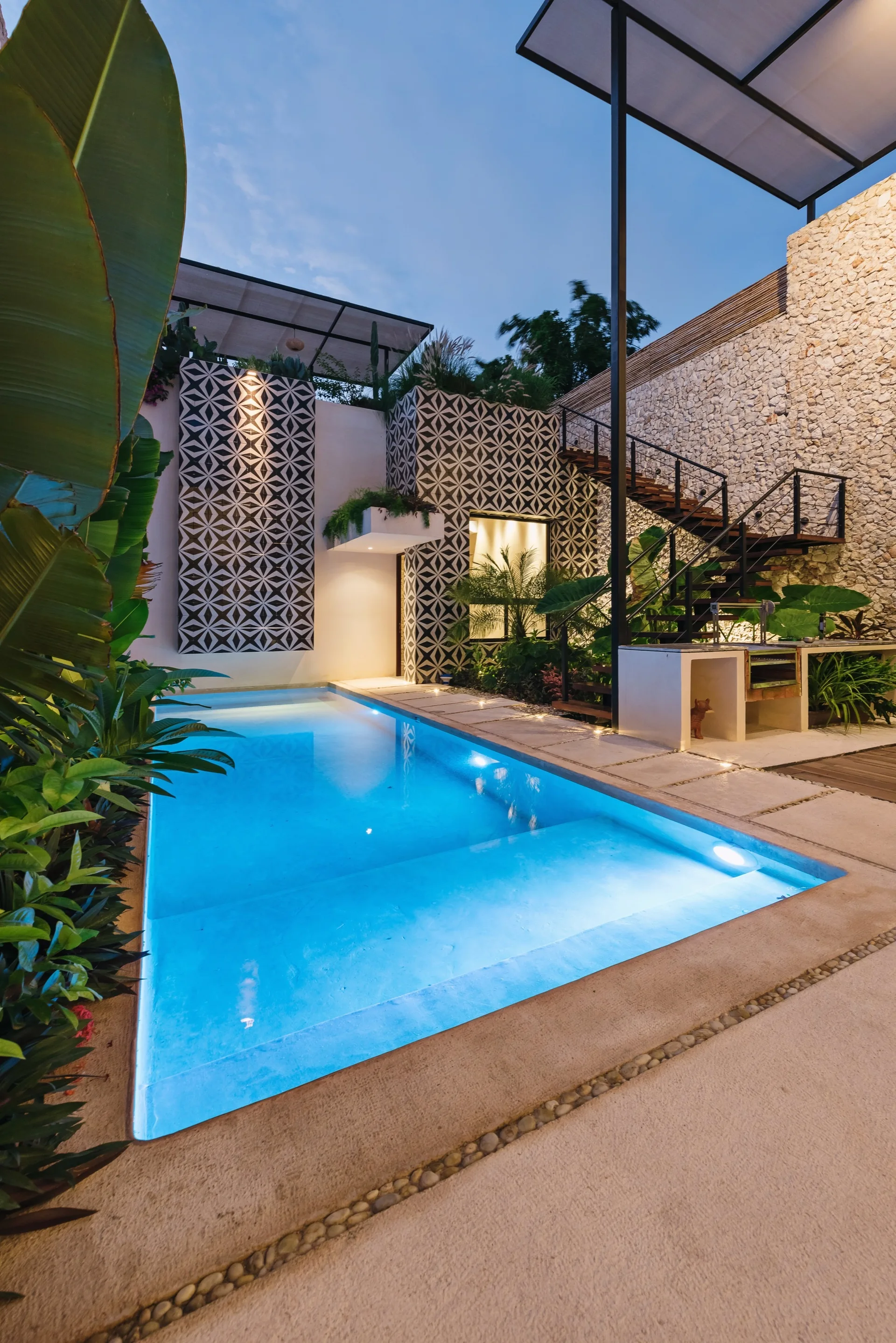 An illuminated outdoor swimming pool in a courtyard featuring bold geometric patterned walls, a metal staircase, and lush tropical plants.