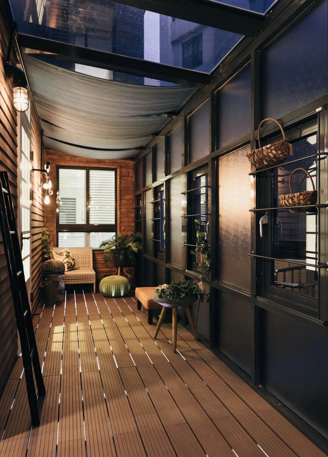 An enclosed patio features horizontal wood decking, a glass panel roof with a fabric shade, and dark metal industrial window frames.