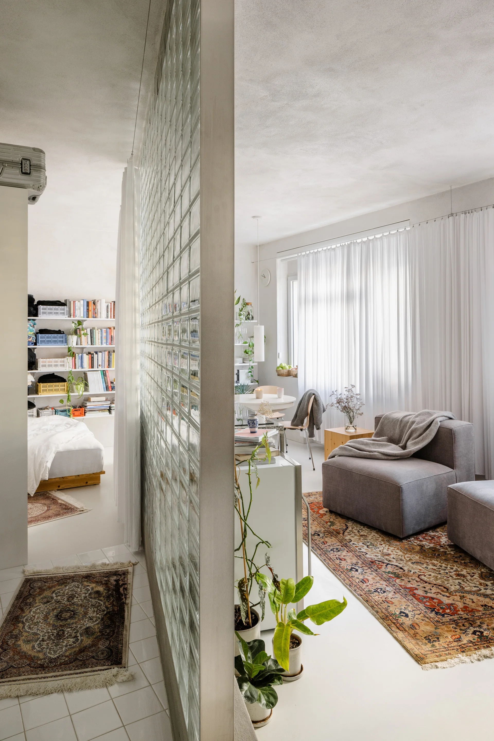 A glass block partition wall divides a living space with grey armchairs from an adjoining sleeping nook in a bright apartment.