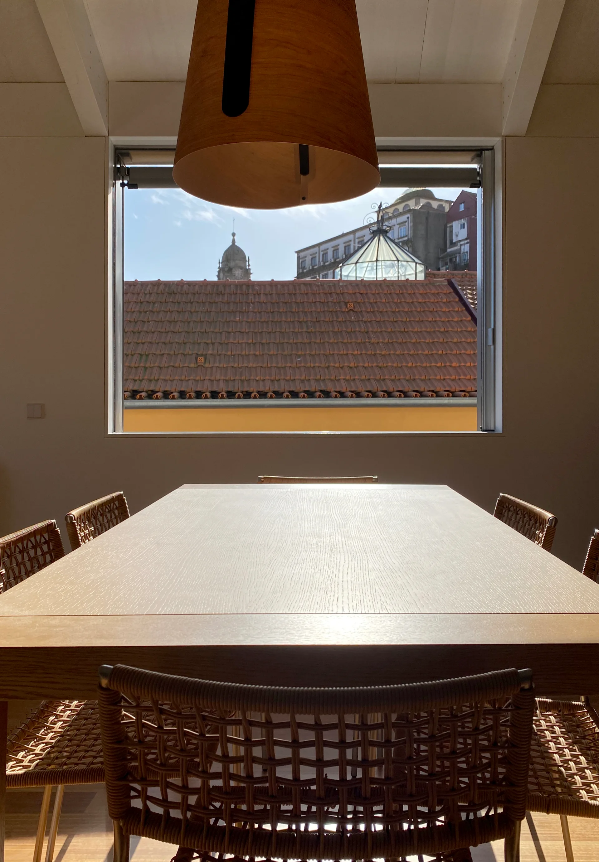 A minimalist dining room features a light wood table surrounded by woven rattan chairs under a large window overlooking red tiled rooftops.