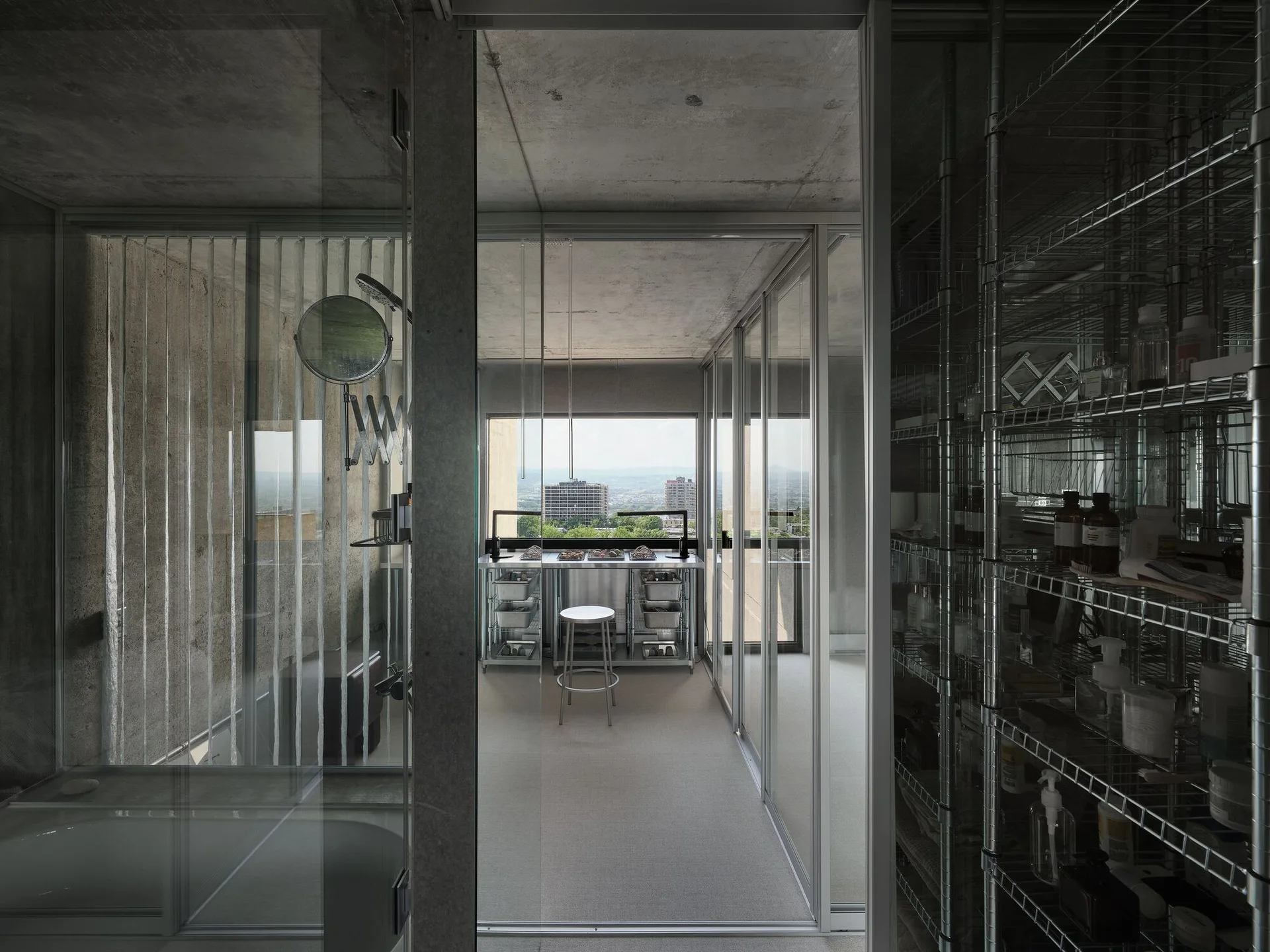 An industrial bathroom features an exposed concrete ceiling, glass partitions, a stainless steel washstand by a window, and chrome wire shelving.