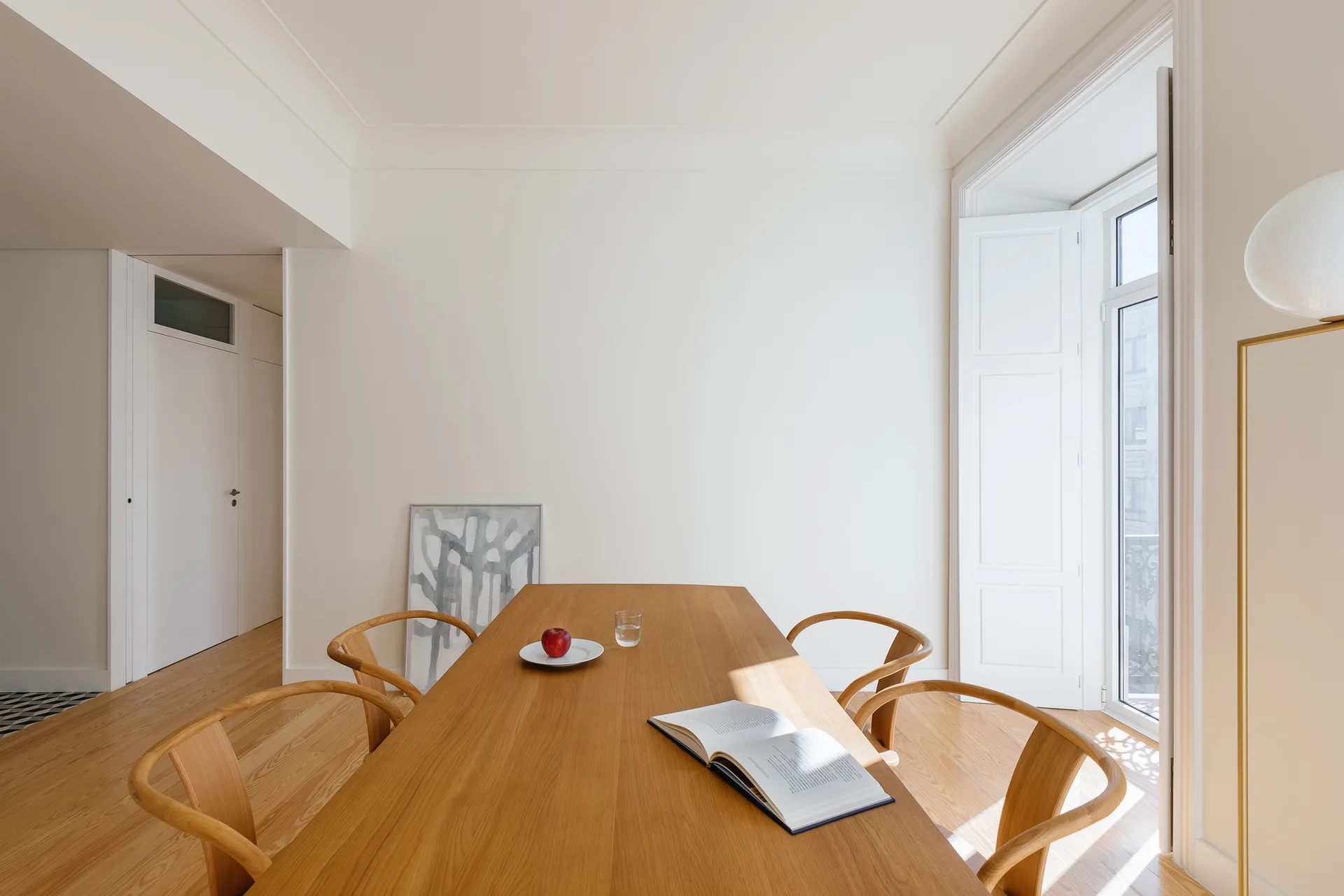 A minimalist dining room features a long wooden table, curved wooden chairs, white walls, and a tall window with white shutters.