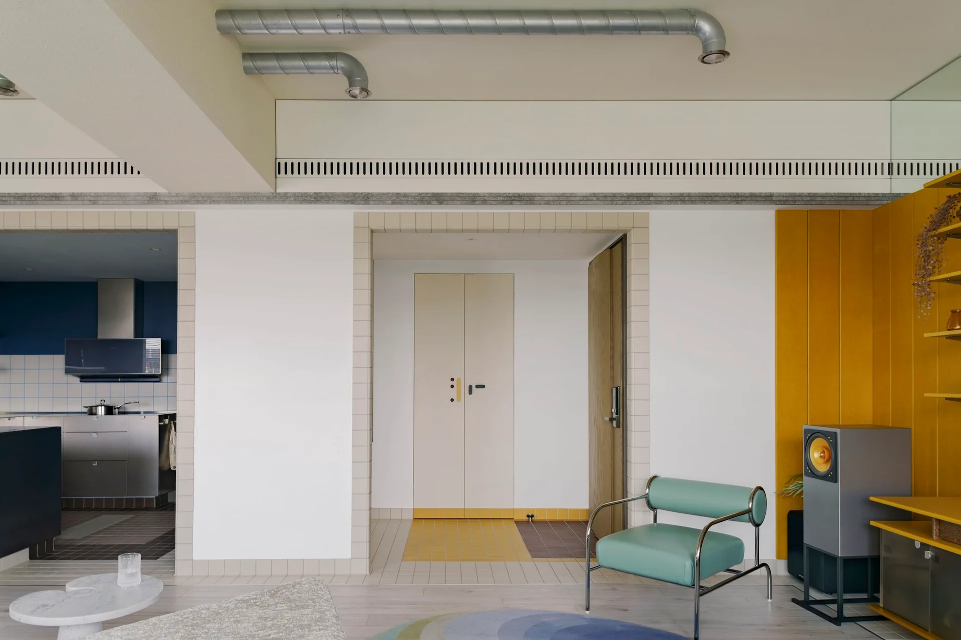 A modern interior room with a mint green armchair, exposed ceiling pipes, yellow built-in shelves, and tiled borders framing wide doorways.