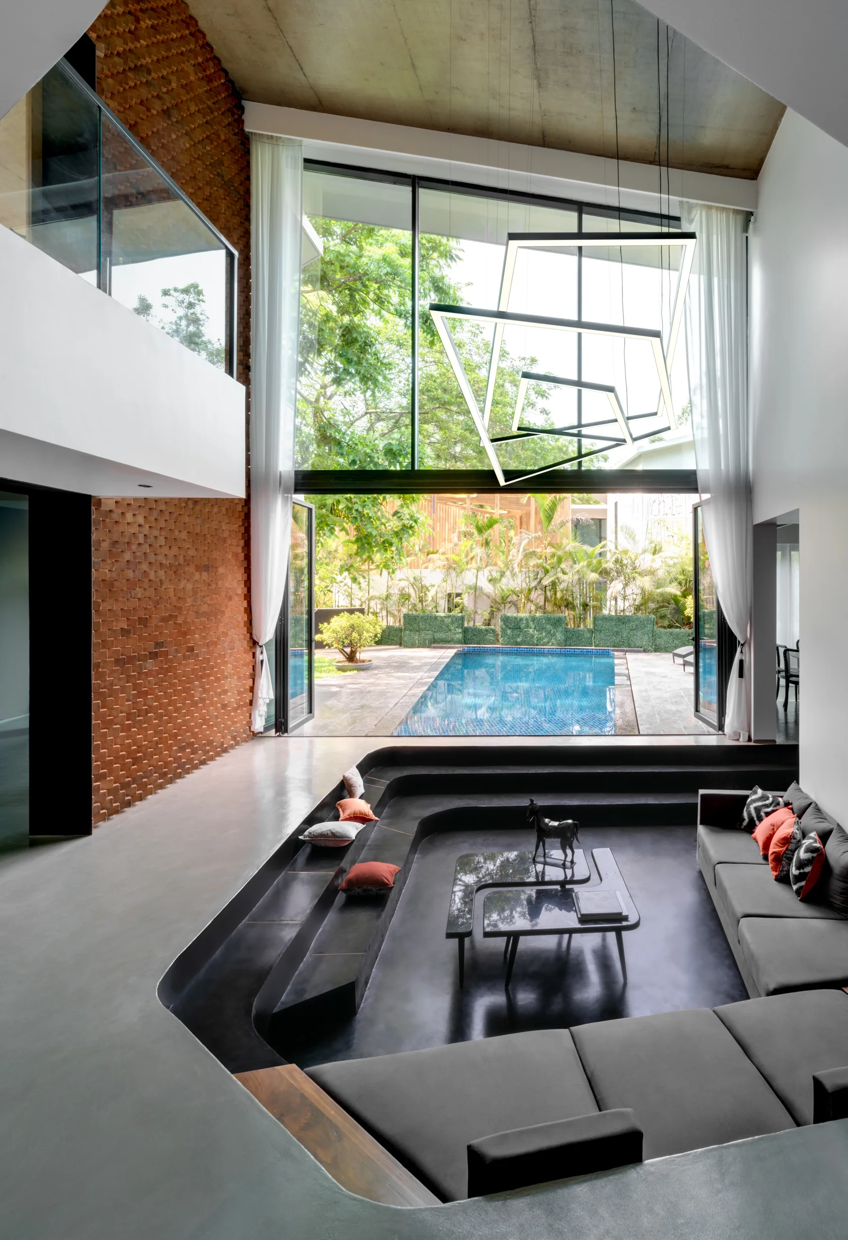 A contemporary sunken living room features a grey sectional sofa, textured brick wall, and high windows overlooking an outdoor pool.
