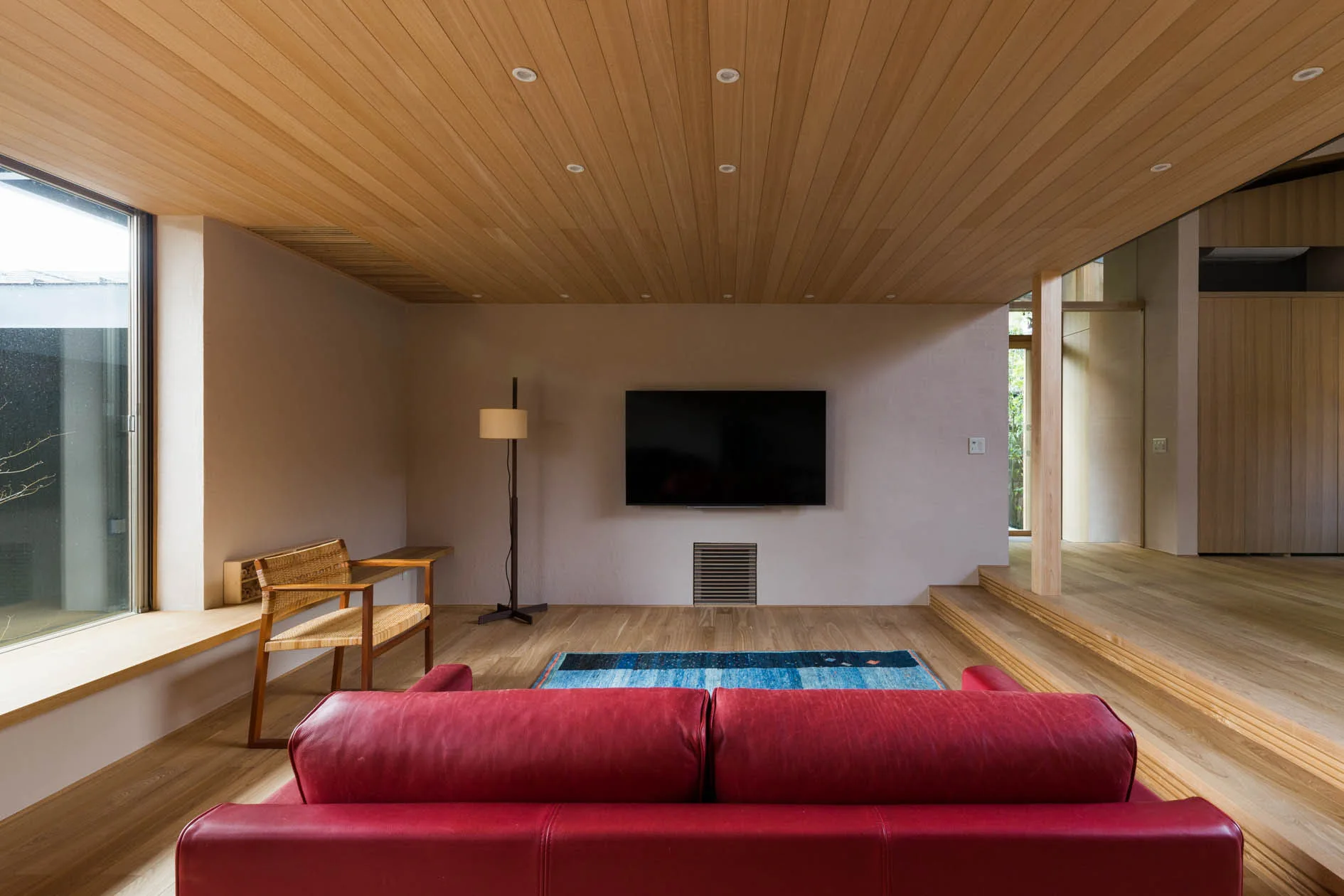 A minimalist living room features a red leather sofa facing a wall-mounted TV under a natural wood plank ceiling.