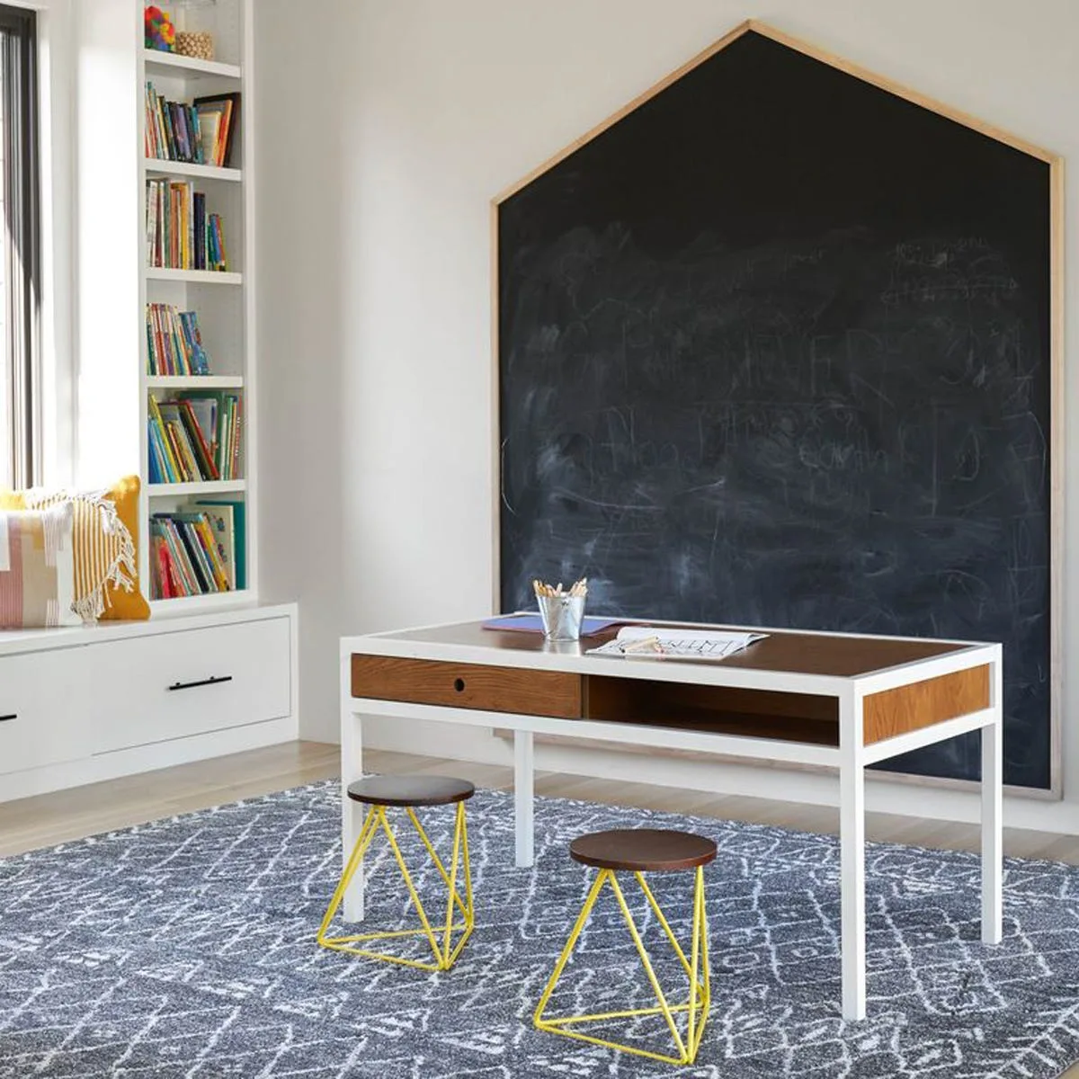 A child's study space features a house-shaped chalkboard, a white and wood desk, two yellow metal stools, and built-in bookshelves.