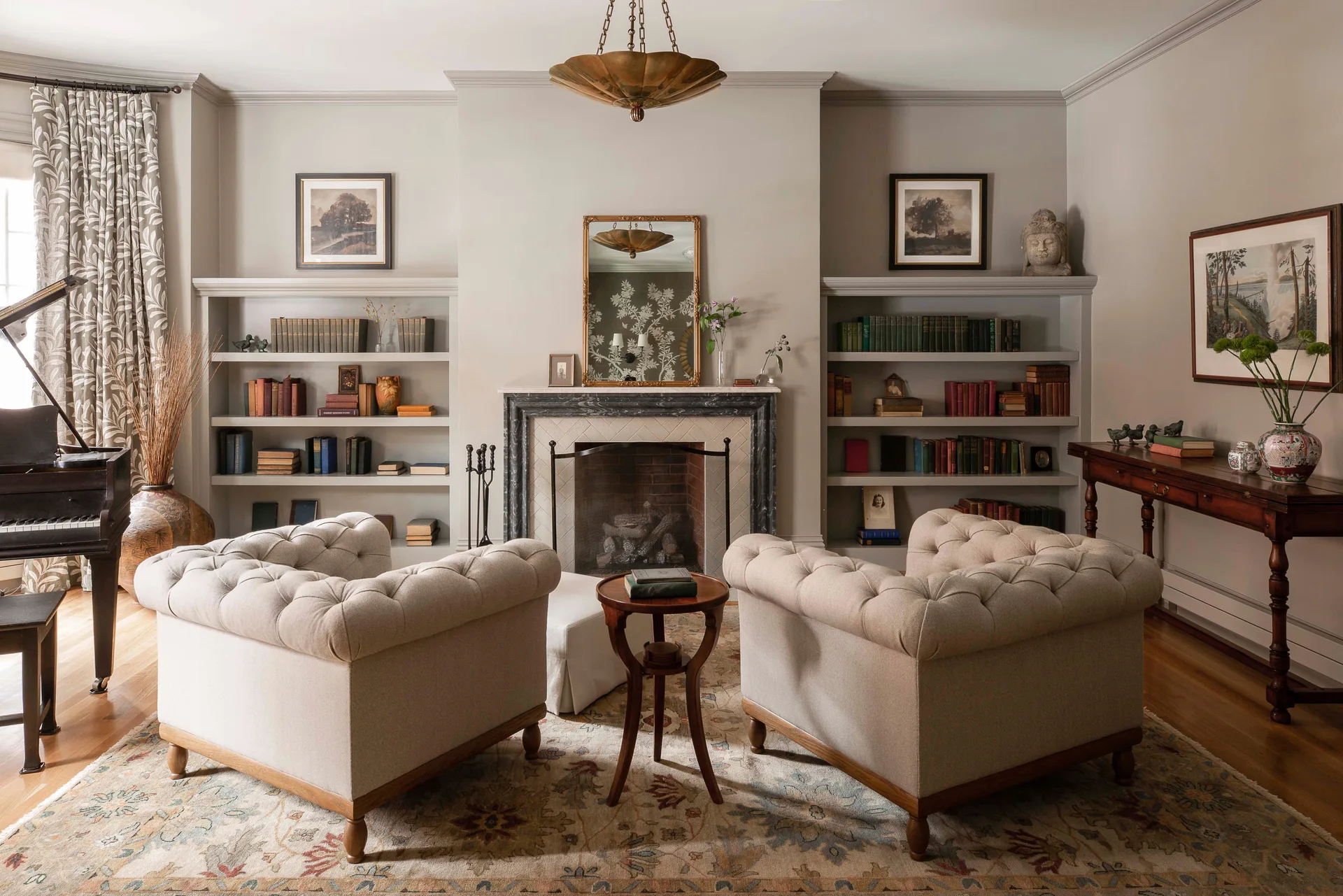 A symmetrical living room features two tufted beige armchairs facing a central fireplace flanked by built-in bookshelves painted to match the walls.