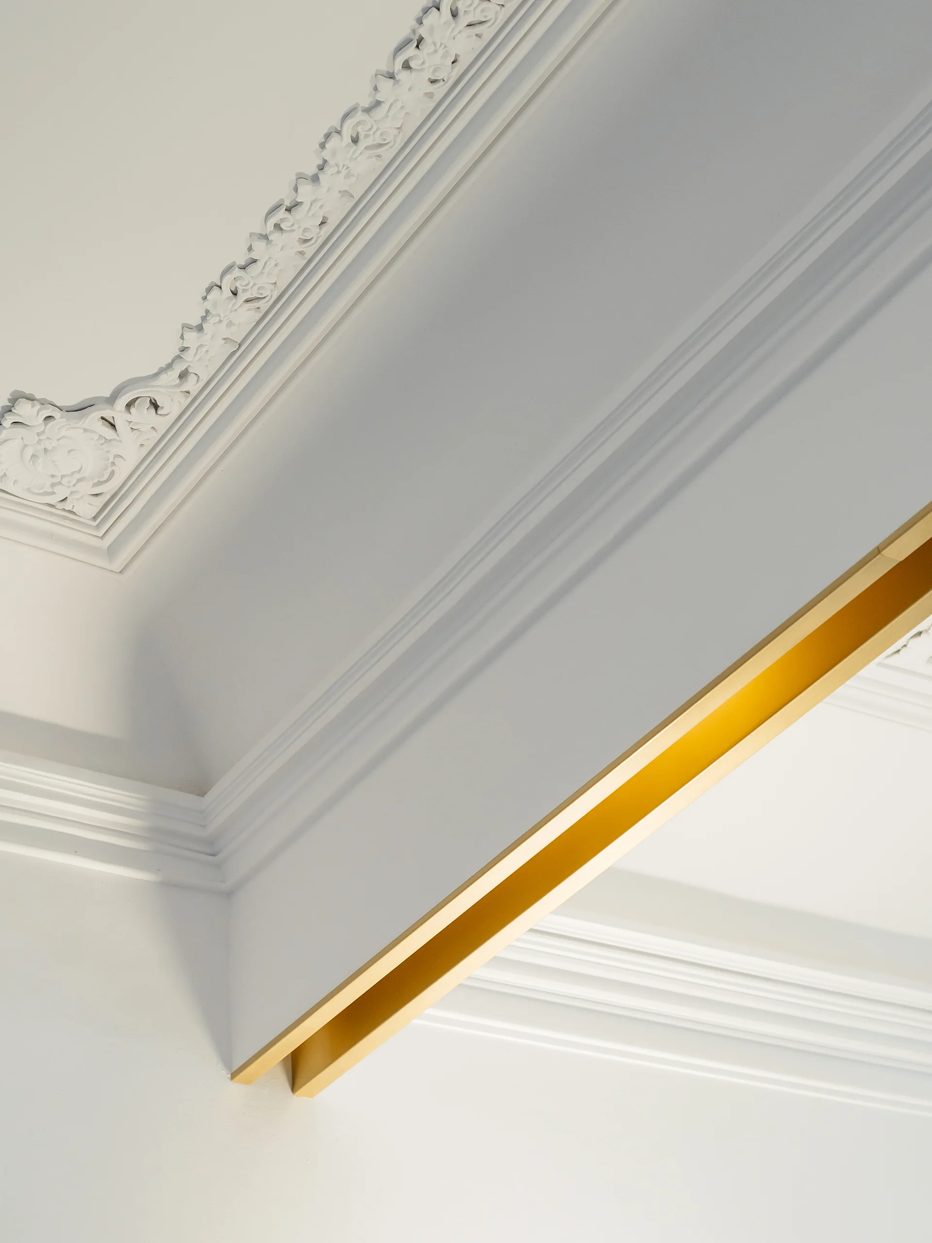 An ornate white plaster crown molding on a ceiling meets a modern gold metal trim accent strip along a clean white wall.