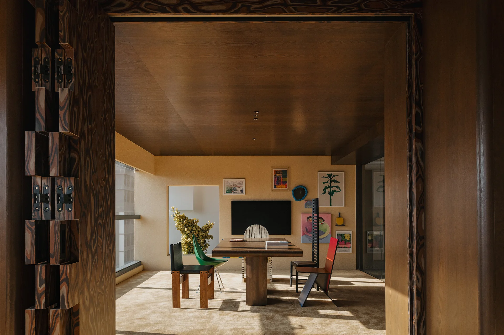 A dining room seen through a patterned wood doorway features eclectic mismatched chairs around a wooden table under a dark wood paneled ceiling.
