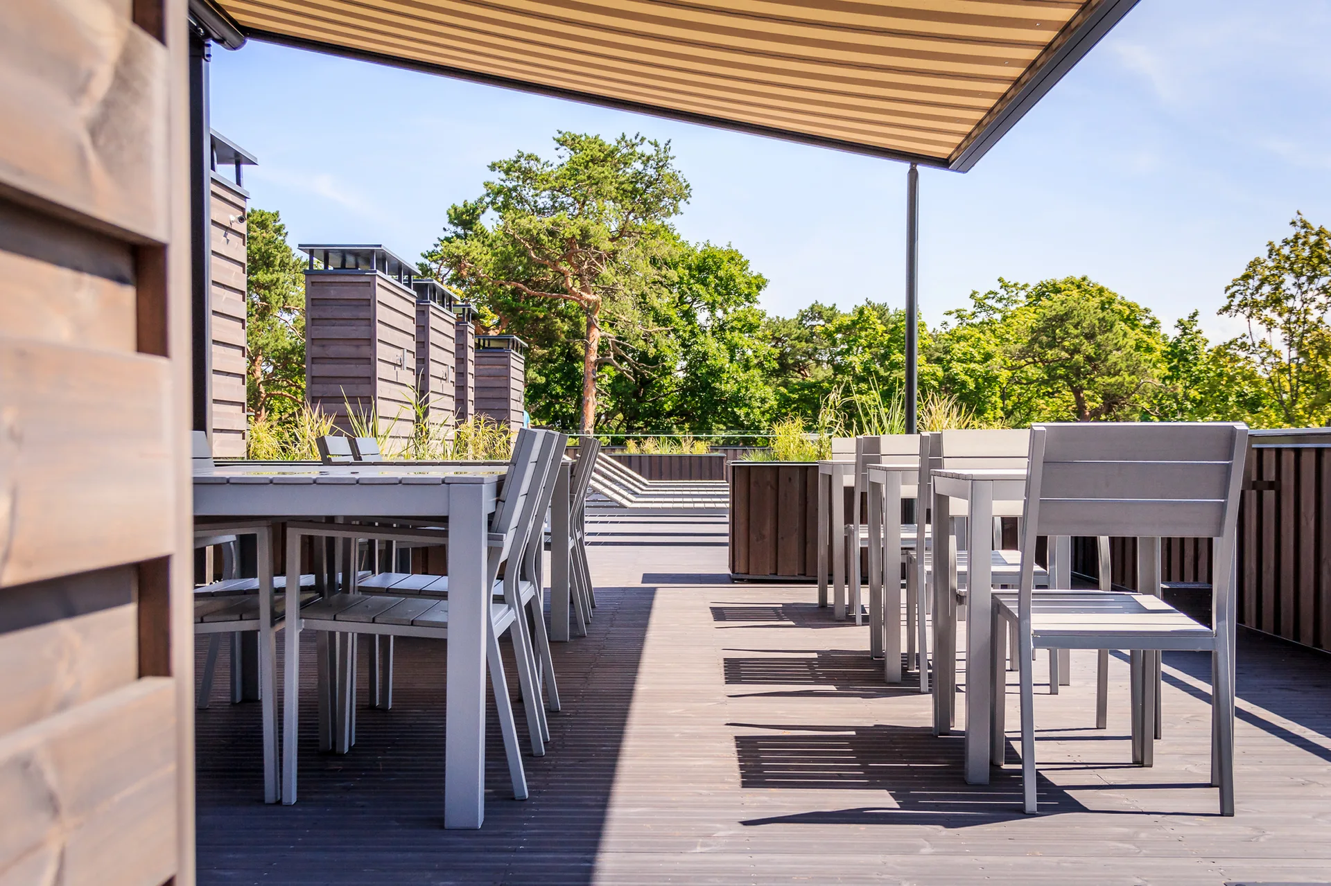 Several gray metal dining tables and chairs stand on a wooden deck under a retractable striped awning.