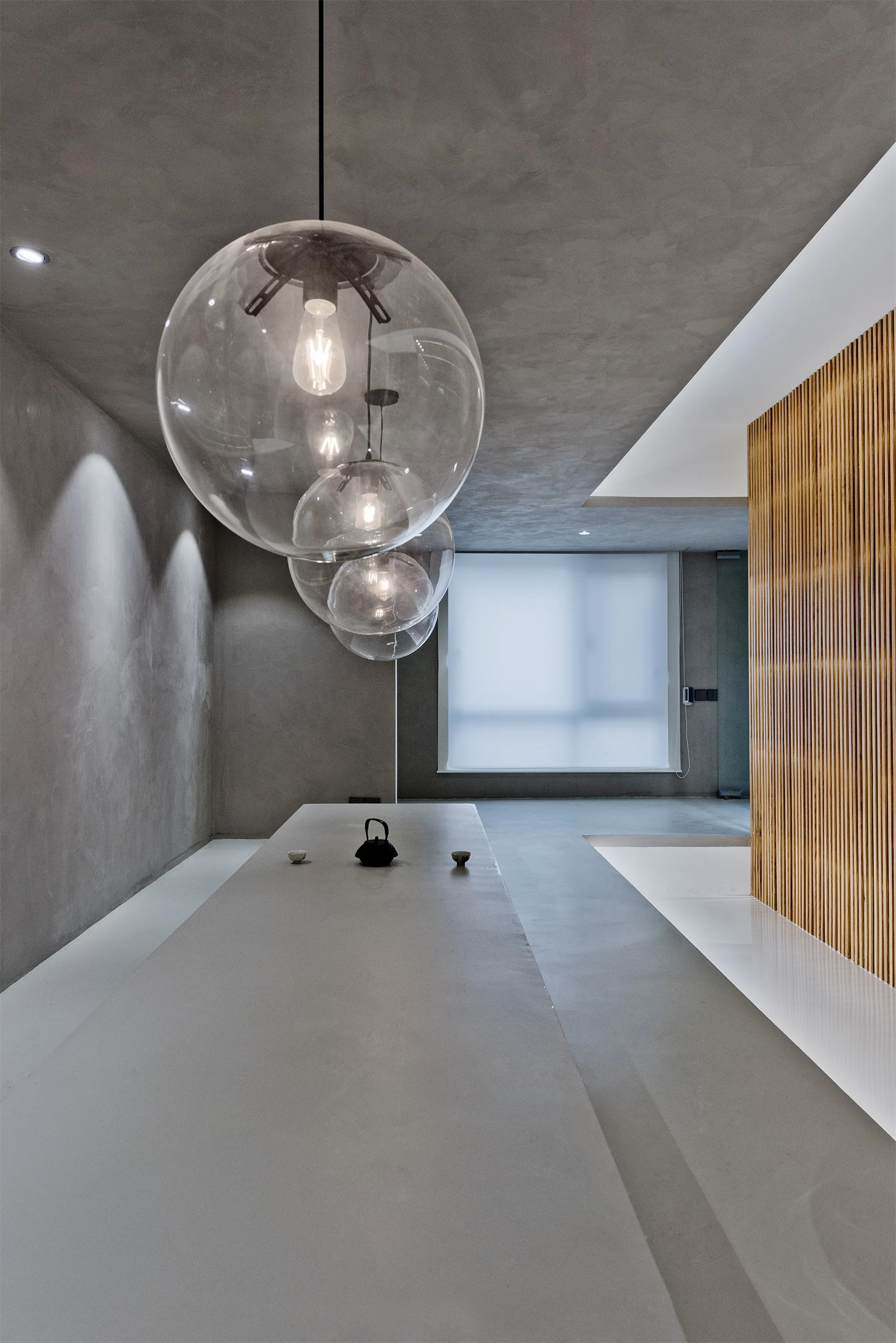 Spherical glass pendant lights hang over a long grey concrete table in a minimalist dining space with a textured wall and vertical wood slats.