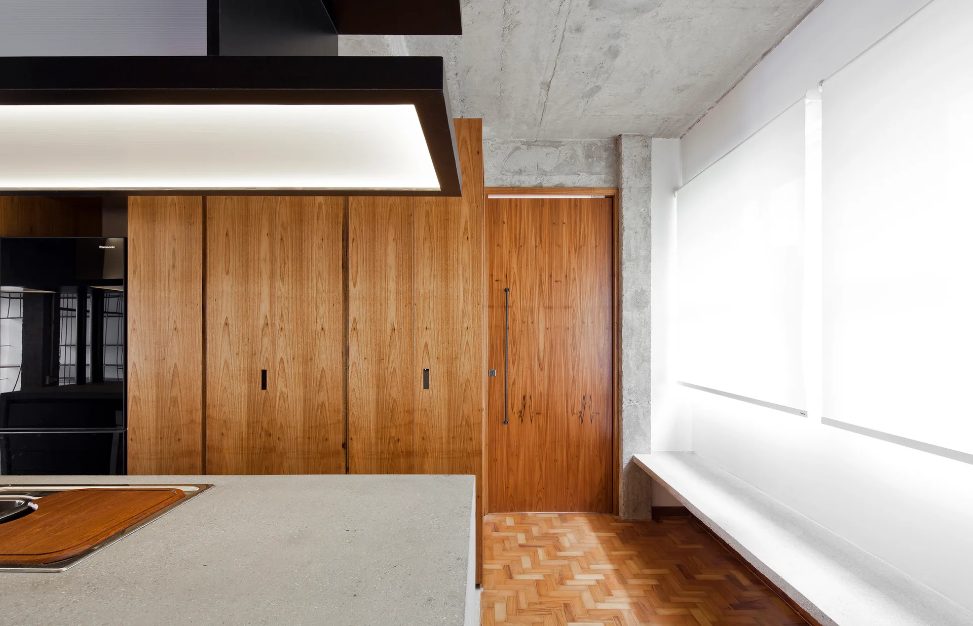 A modern kitchen features full-height wood cabinetry, a flush door, a stone island, and an exposed raw concrete ceiling.