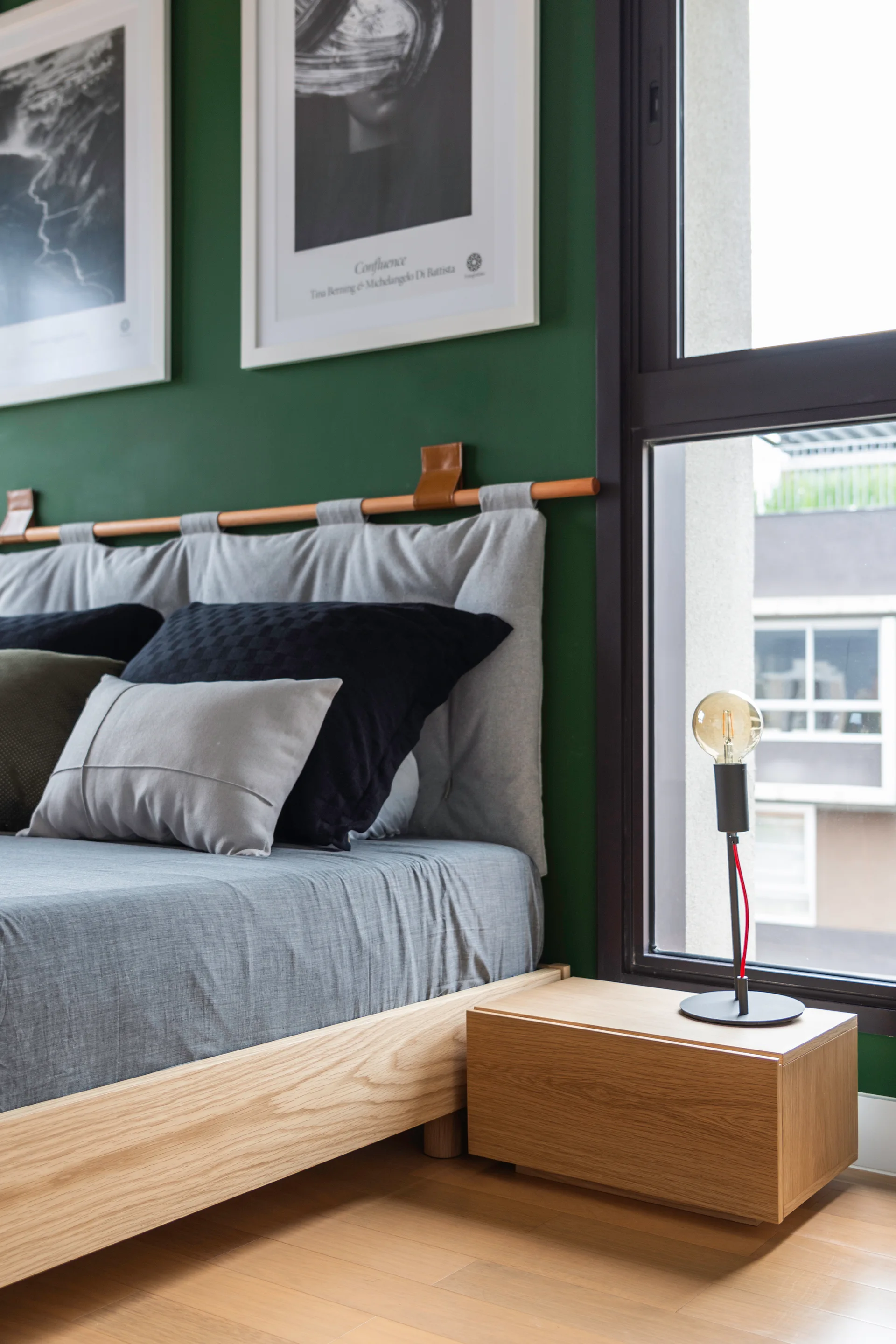 A low wooden bed frame with a gray fabric headboard sits against a deep green accent wall next to a minimalist wooden nightstand and window.