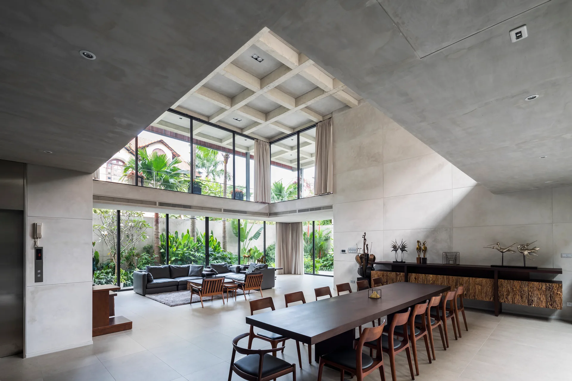 A modern open-plan dining room features a long wooden table, concrete walls, a coffered ceiling, and glass windows overlooking a tropical garden.