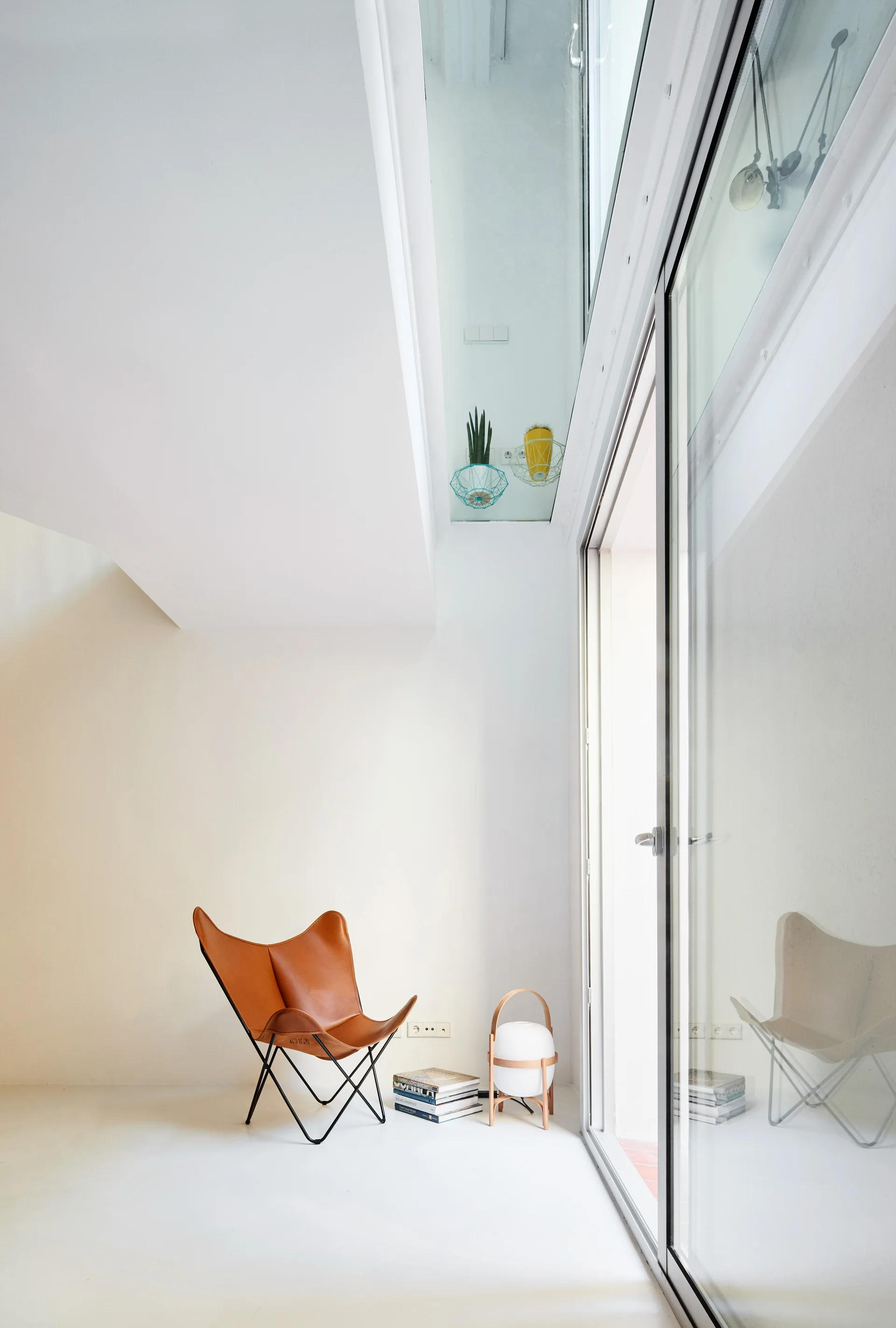 A minimalist white room features a tan leather butterfly chair, a stack of books, a portable floor lamp, and a large sliding glass door.
