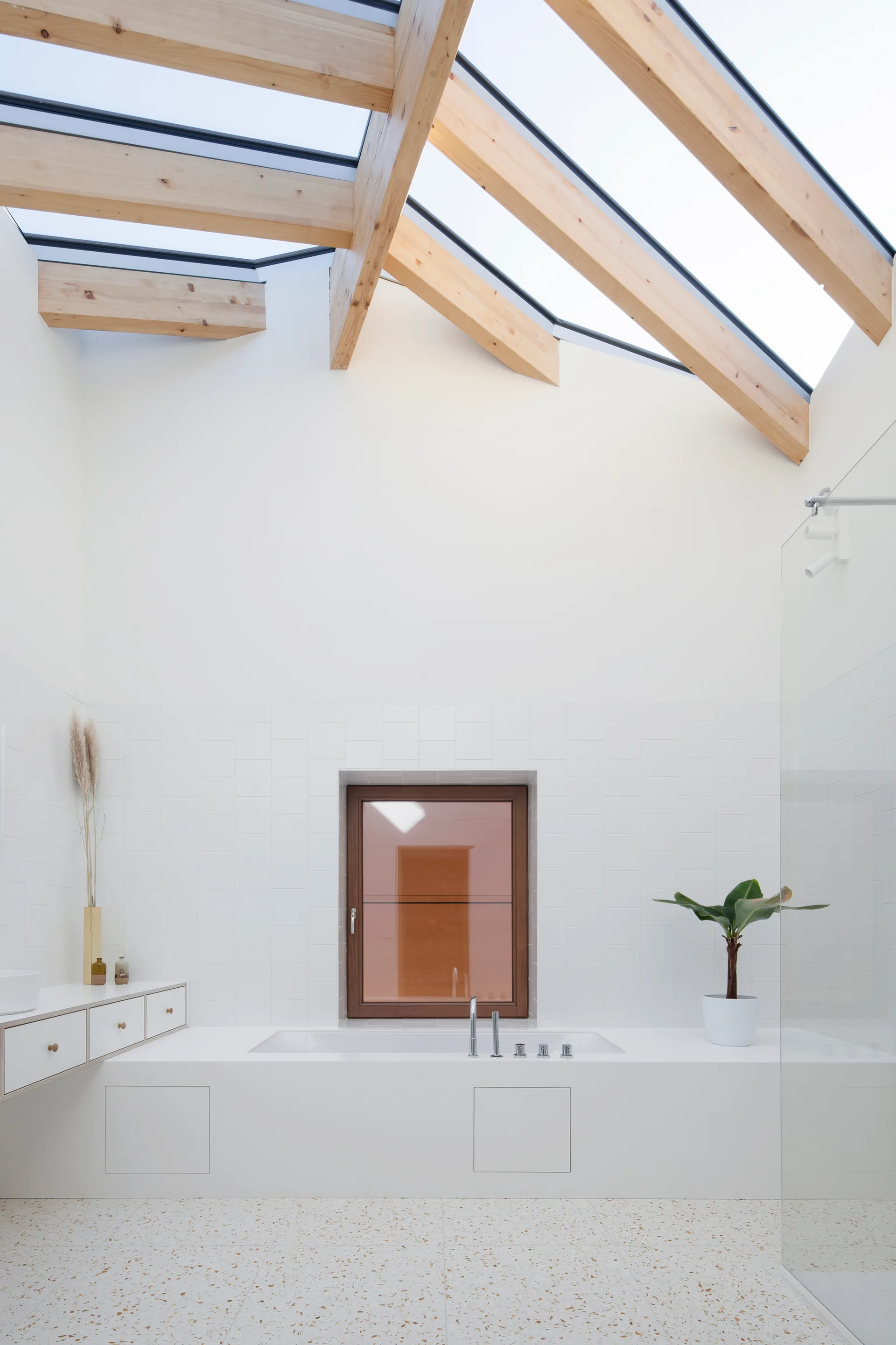 A minimalist white bathroom features a built-in tub under a large sloped skylight with exposed light wood beams and a terrazzo-tiled floor.