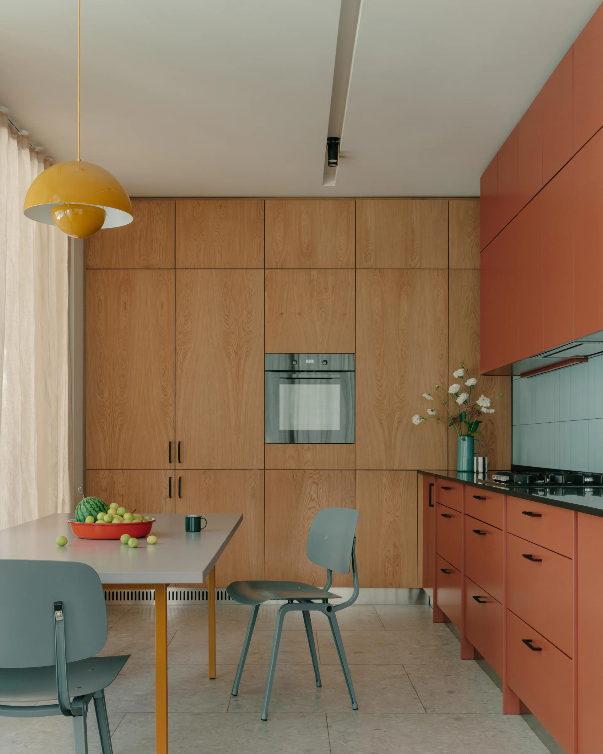 A modern kitchen featuring tall wood veneer cabinets, rust red base units, a grey dining table with matching chairs, and a yellow pendant light.