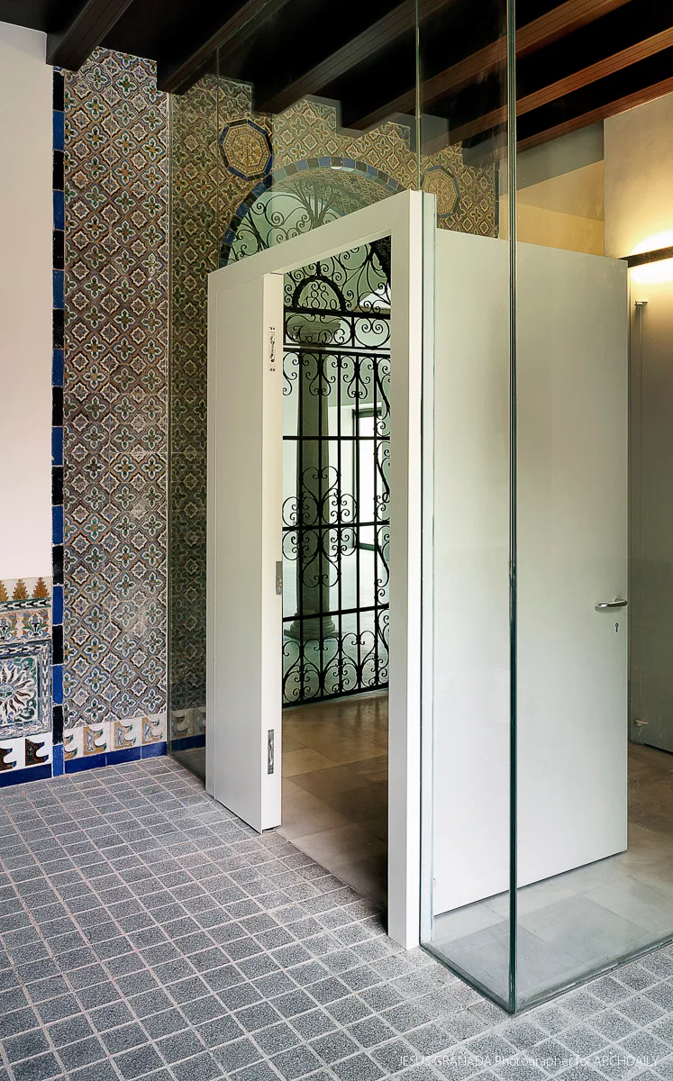 A modern frameless glass wall and white door stand in front of a historic wall covered in intricate geometric blue and gold ceramic tiles.