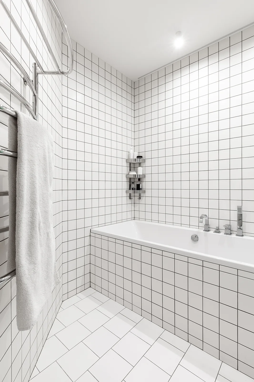 A minimalist white bathroom featuring grid-tiled walls, a built-in bathtub with chrome fixtures, and a heated towel rail.
