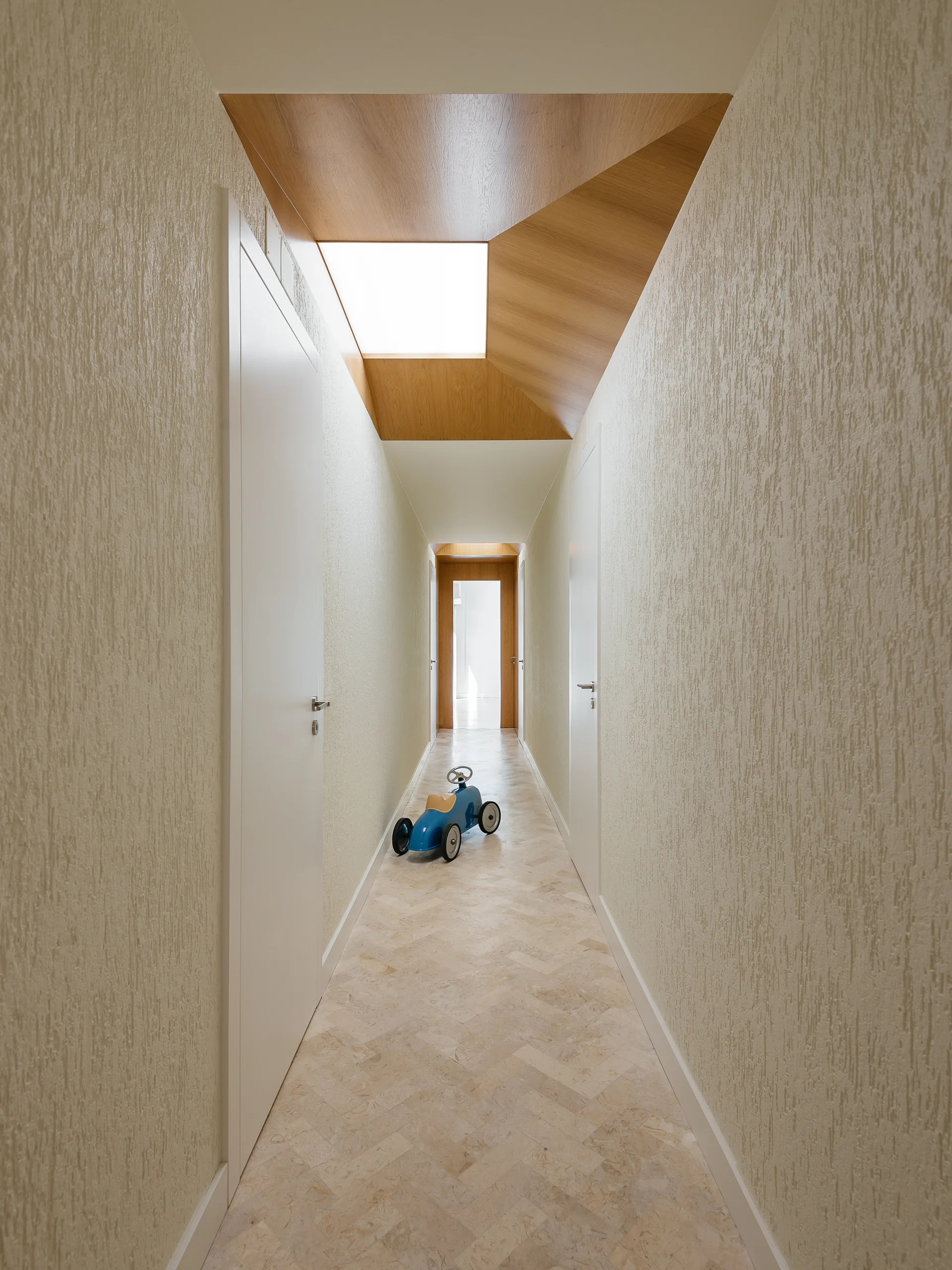 A long residential hallway featuring textured beige walls, flush white doors, a herringbone stone tile floor, and a geometric wood-clad skylight.
