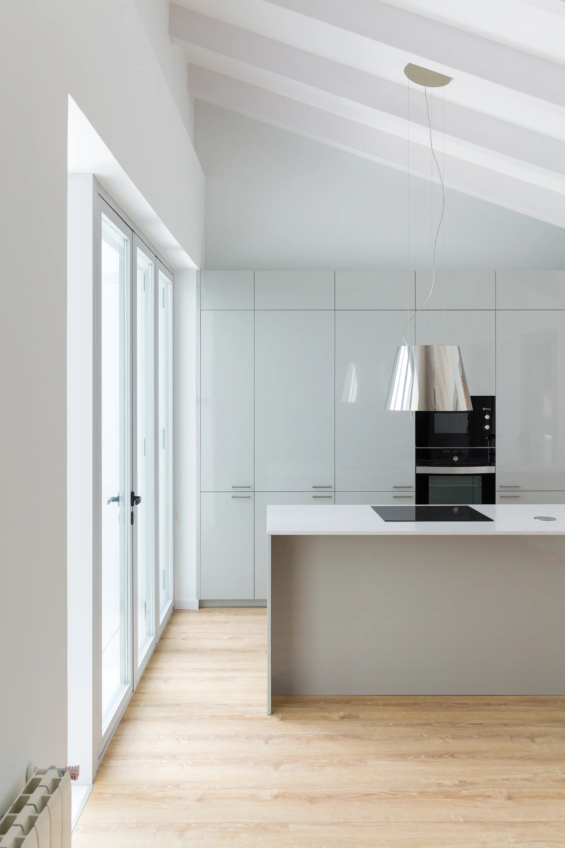 A minimalist kitchen featuring light grey gloss cabinetry, a central island, wood floors, and a vaulted beamed ceiling next to glass doors.