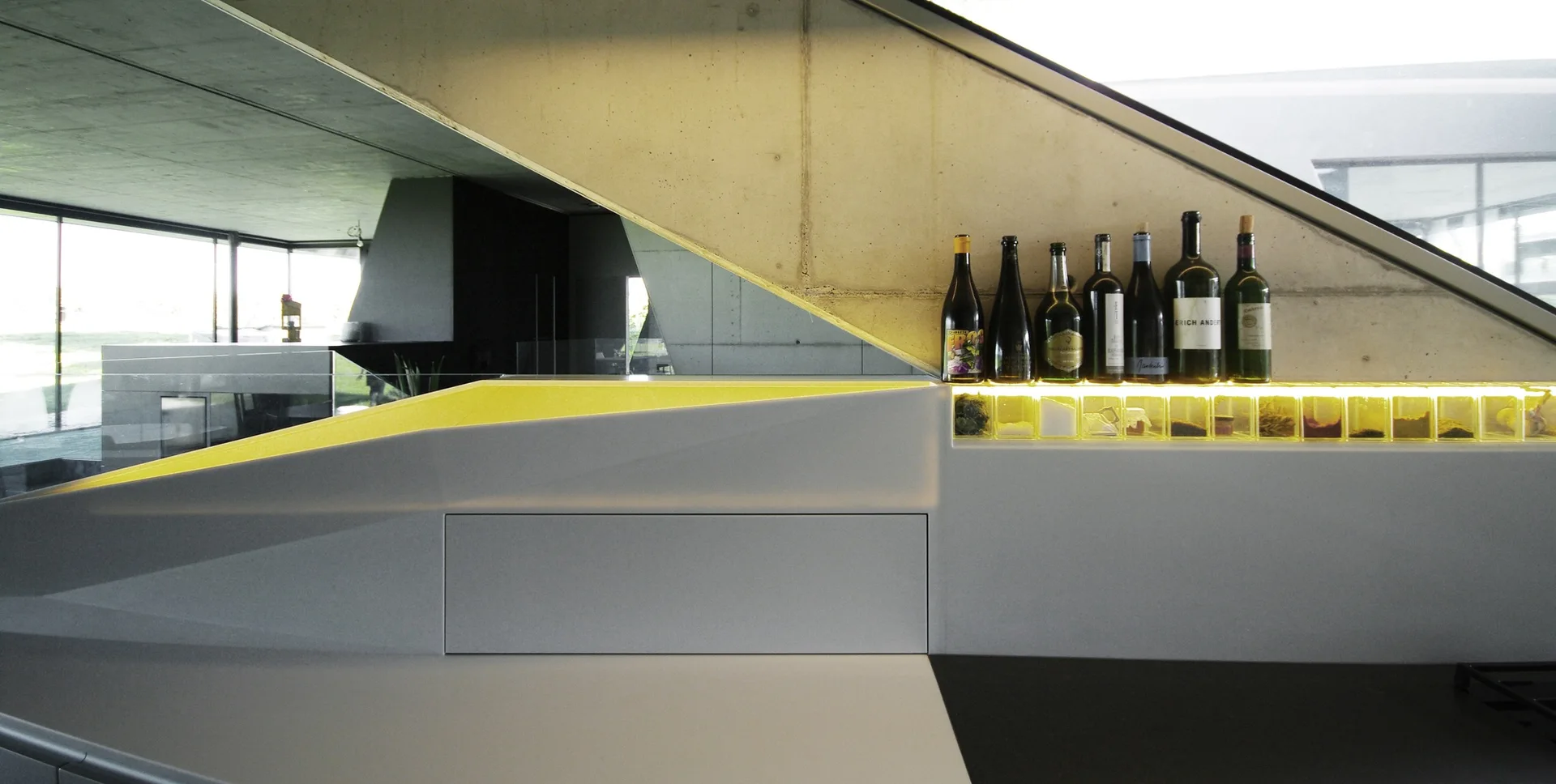 An angular white kitchen counter with backlit yellow glass compartments underneath wine bottles, set against a sloped concrete wall.