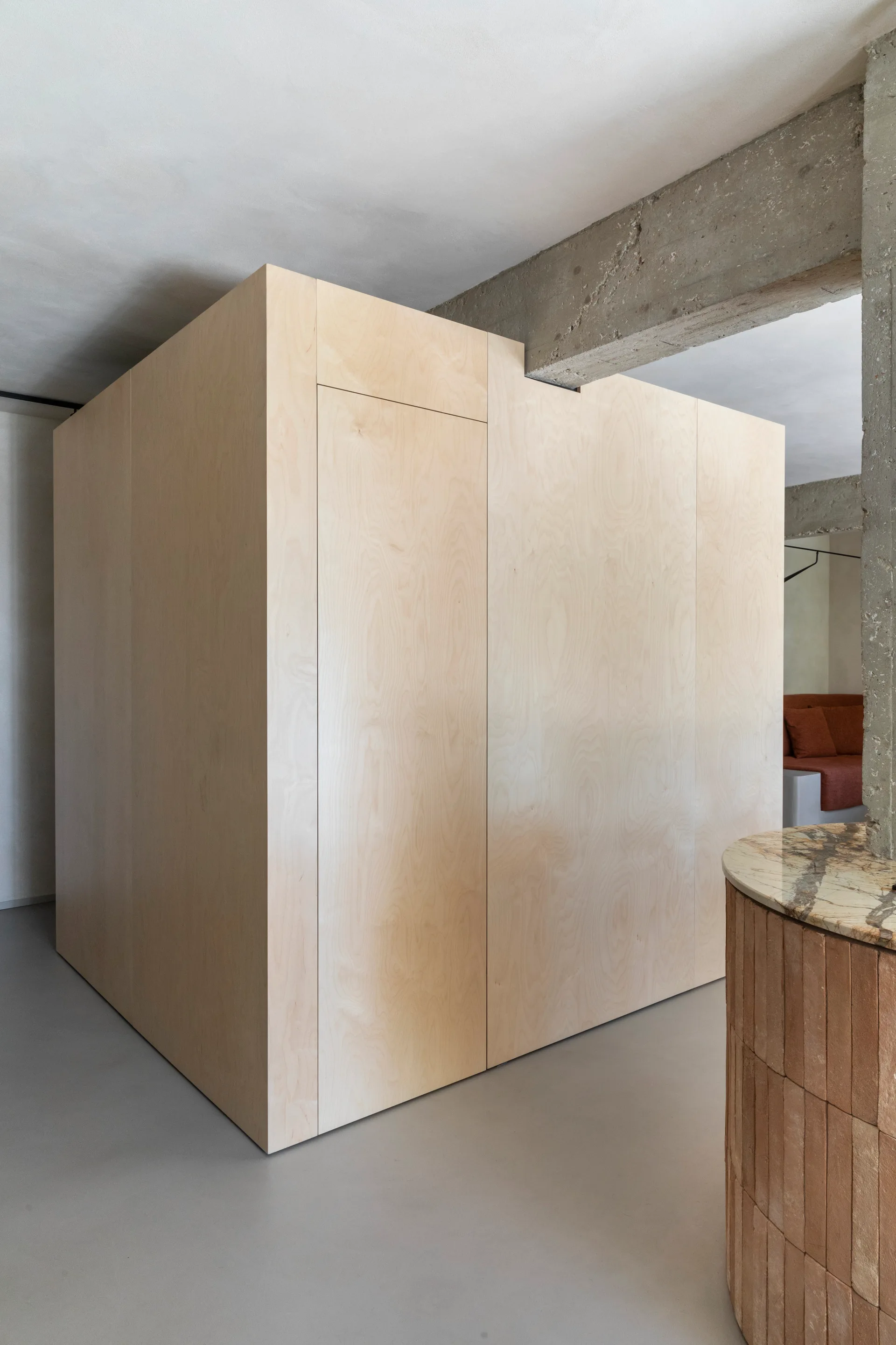 A large minimalist plywood storage structure stands on a grey floor beneath an exposed concrete ceiling beam in an industrial loft.