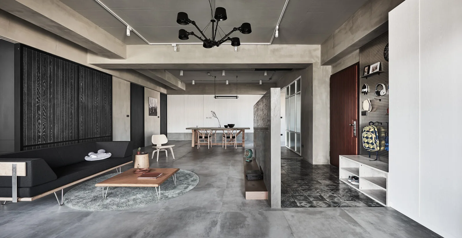 An industrial living room features a concrete partition wall, a black contemporary sofa on a round rug, and an entryway pegboard.
