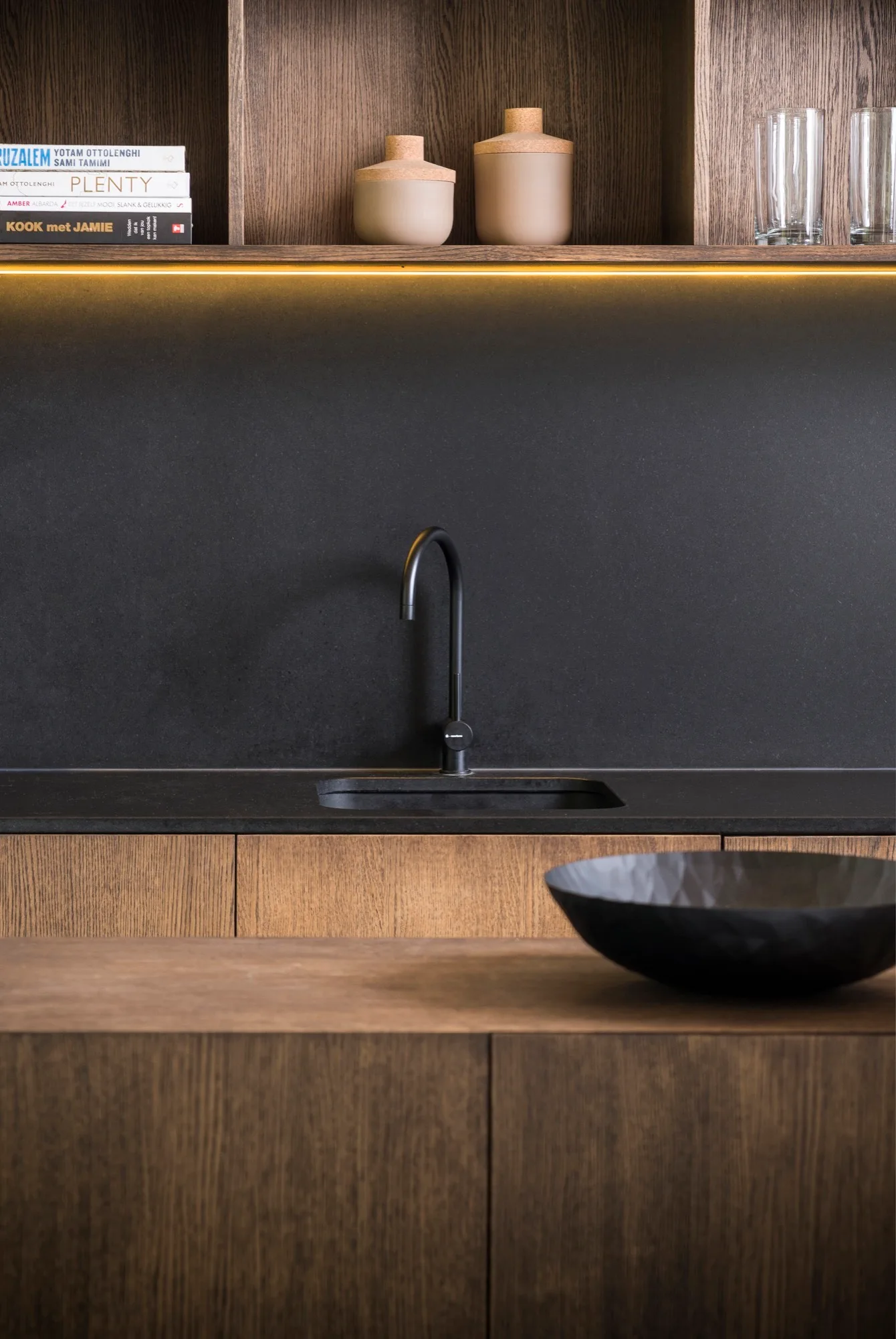 A modern kitchen sink features a matte black faucet and backsplash paired with warm wood cabinetry, open shelving, and integrated lighting.