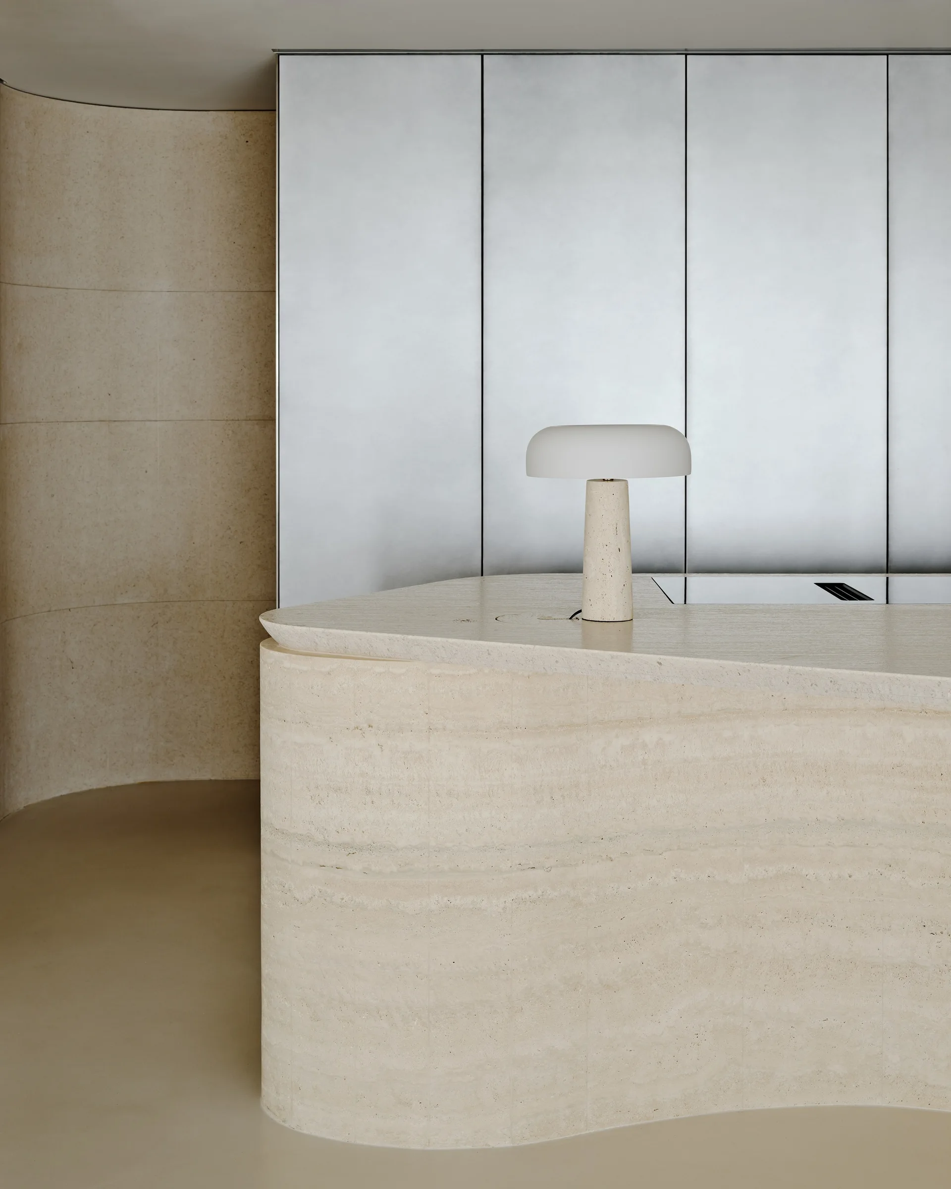 A curved travertine stone kitchen island with a small dome lamp sits in front of tall brushed metal cabinet panels.