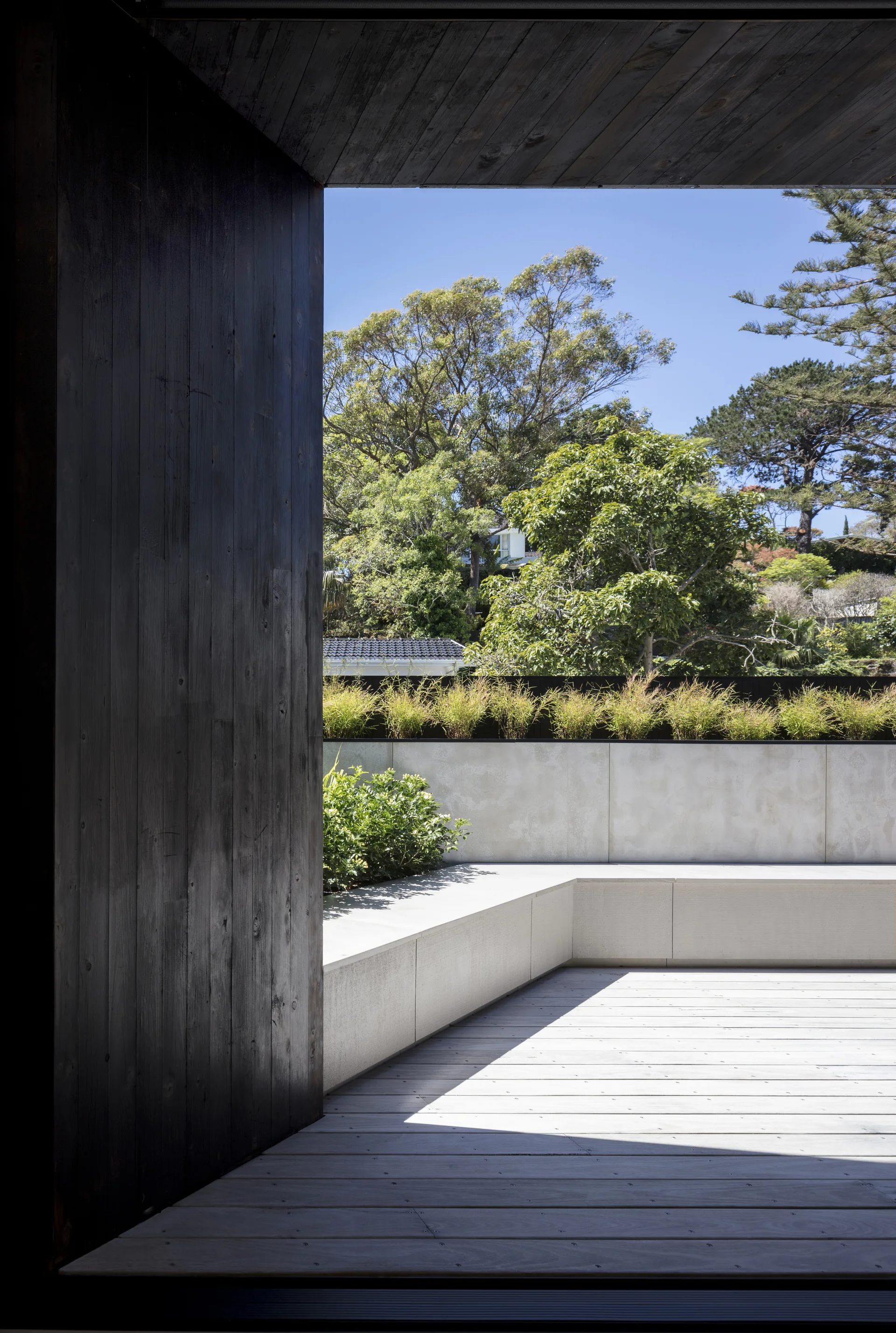 A modern outdoor patio features a built-in concrete bench, light wood decking, and a concrete retaining wall under a dark wood ceiling.