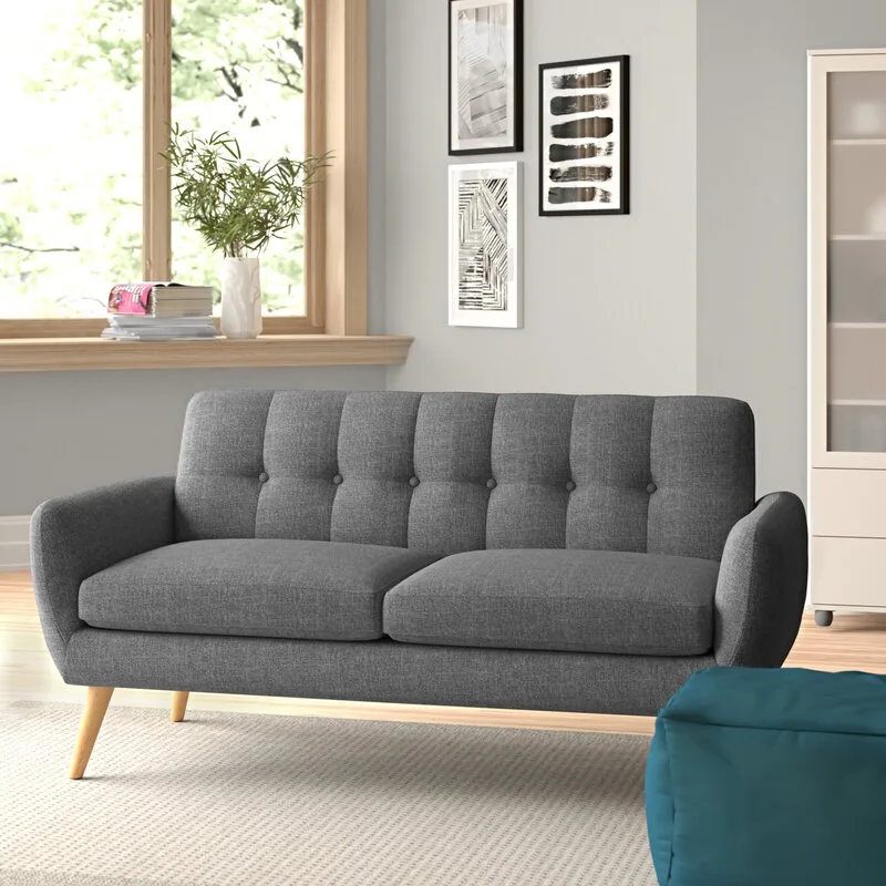 A gray tufted sofa with tapered wood legs sits on a textured rug in a bright room with framed art and a window.