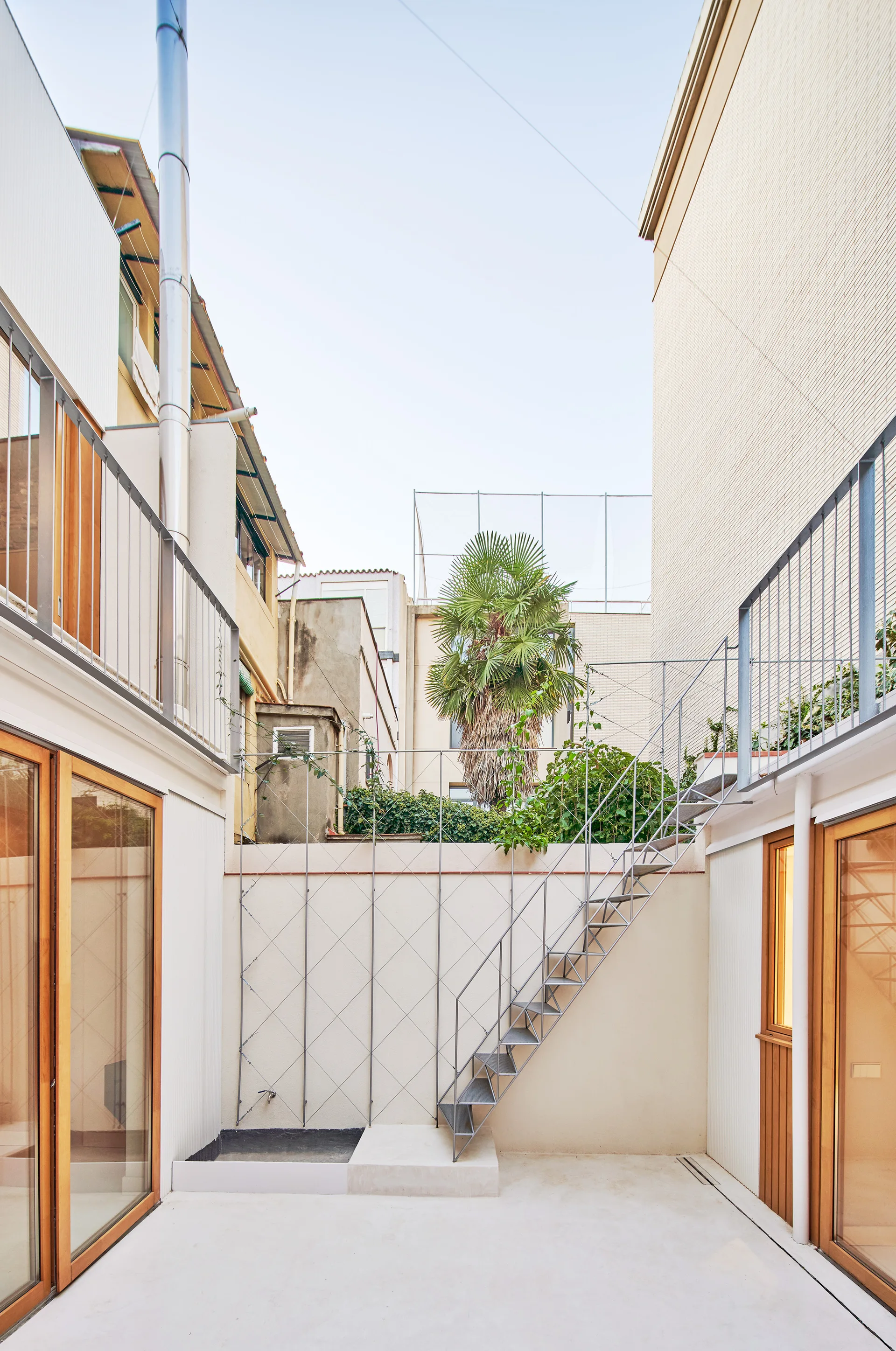 An urban courtyard with off-white walls, wood-framed glass doors, an open-riser metal staircase, and a diamond-patterned wall trellis.