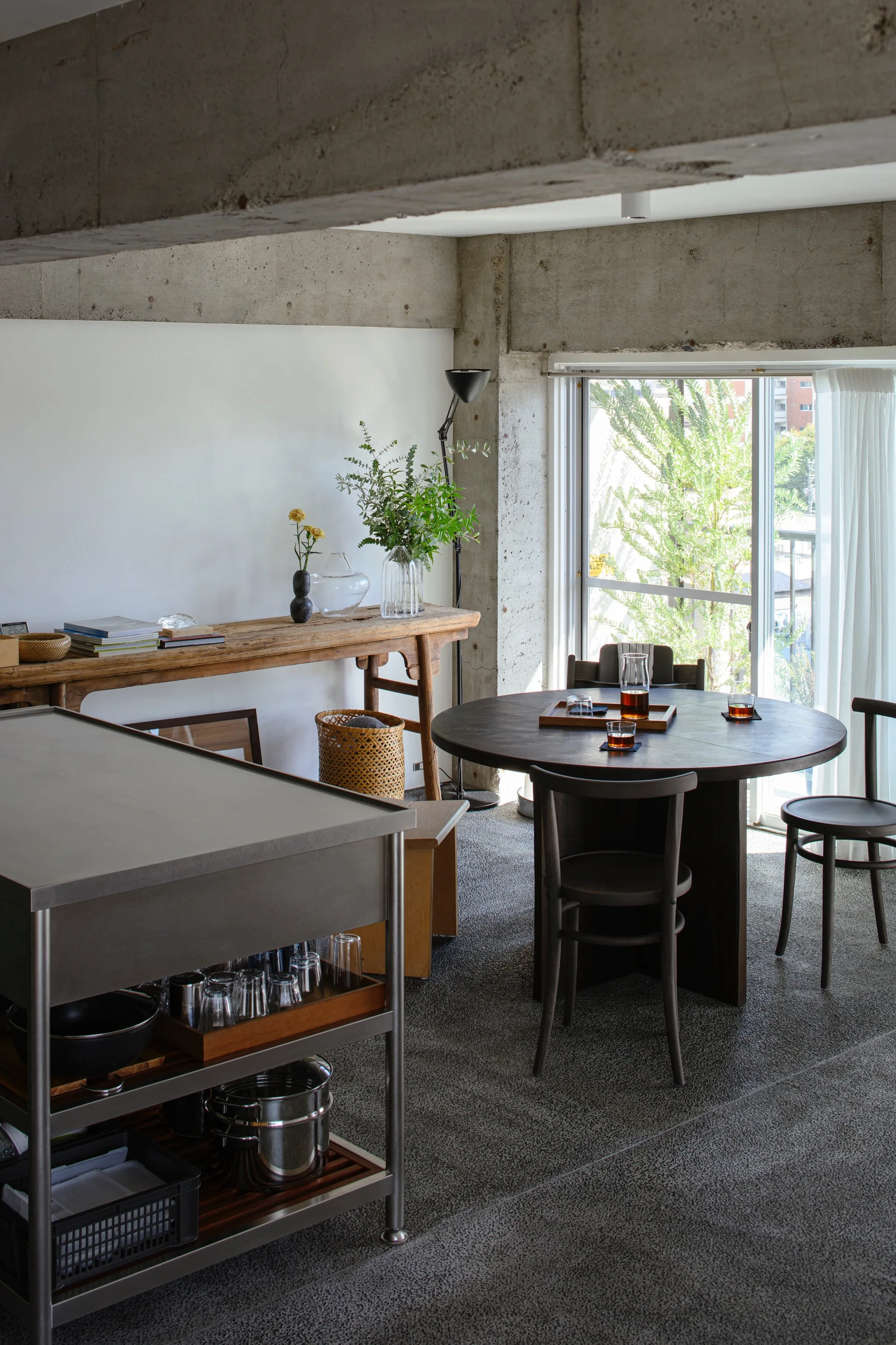 A minimalist dining space featuring a round dark wood table, matching chairs, an exposed concrete beam, and a rustic wooden console table.