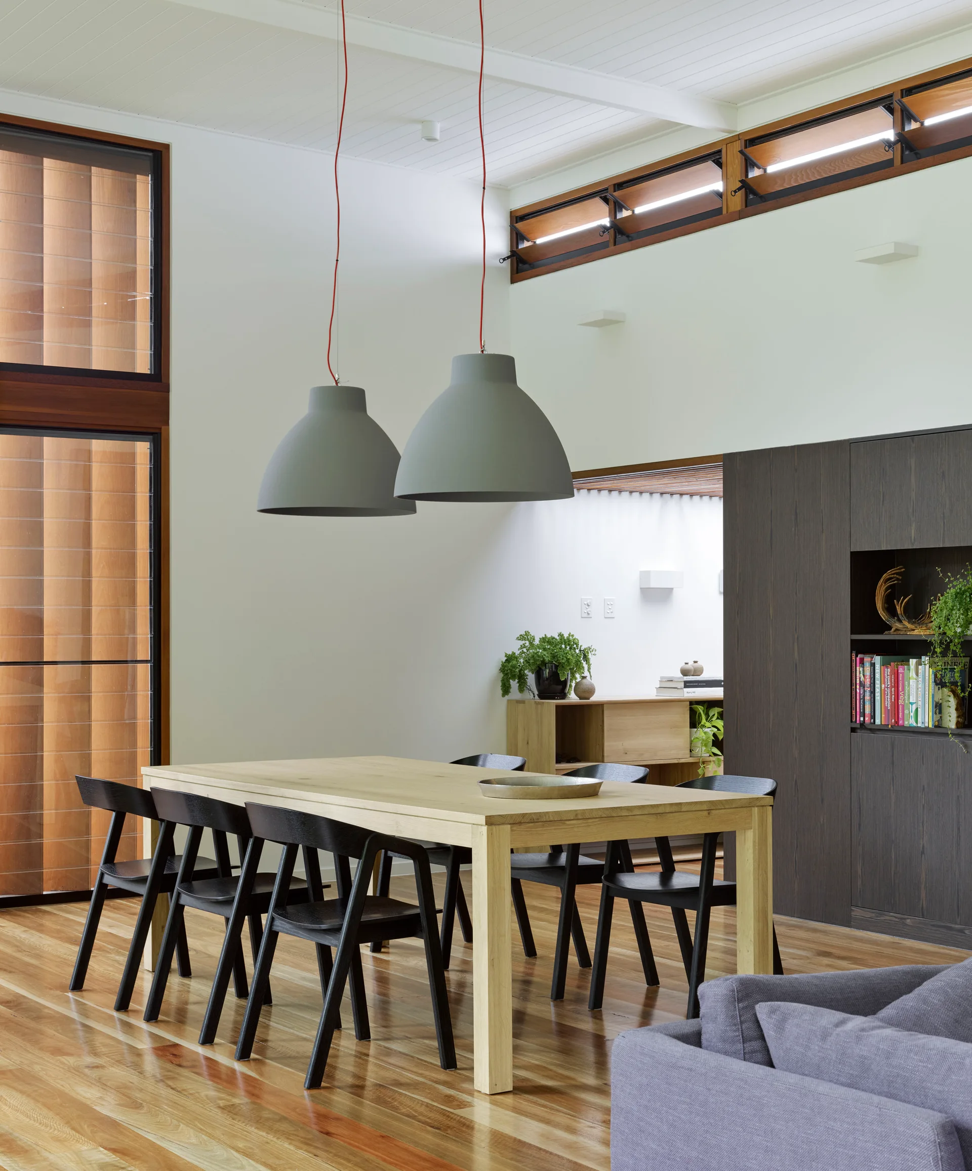 A modern dining room features a light wood table surrounded by black chairs beneath two large grey dome pendant lights, with wood-framed windows.