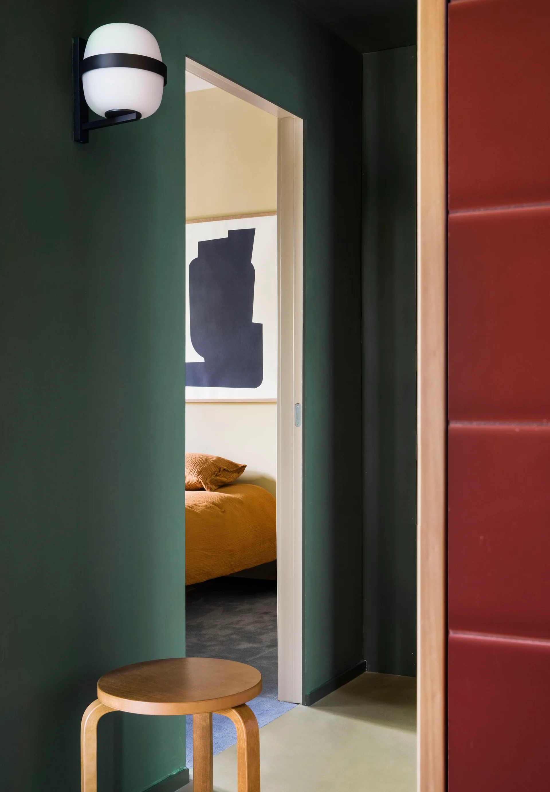 A view through a dark green doorway into a bedroom featuring ochre bedding and a large black and white abstract print on a light wall.