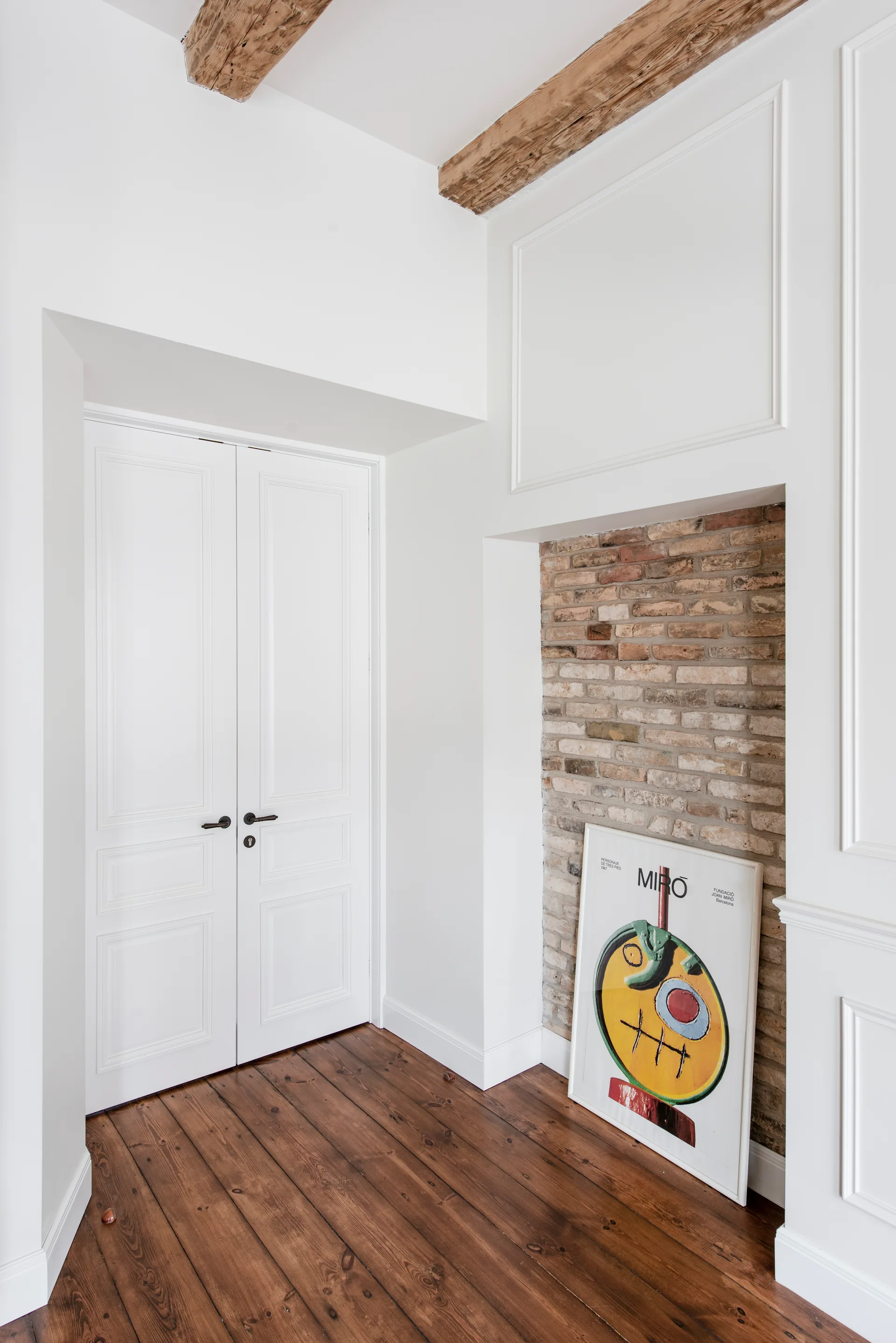 A bright hallway features white paneled double doors, wood flooring, and a recessed exposed brick wall section where a framed art print rests on the floor.