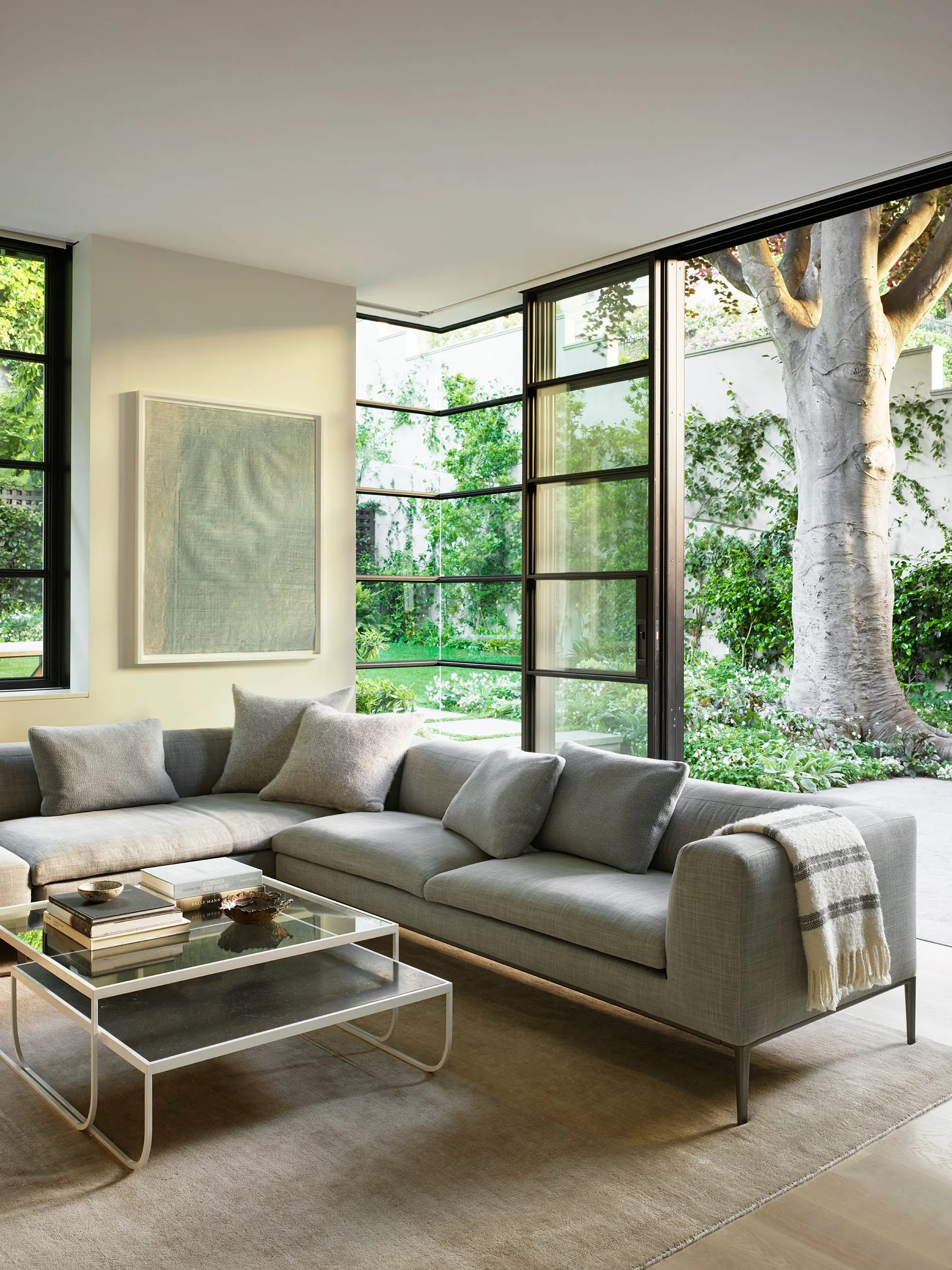 A modern grey sectional sofa and glass coffee table face floor-to-ceiling corner windows overlooking a green garden.