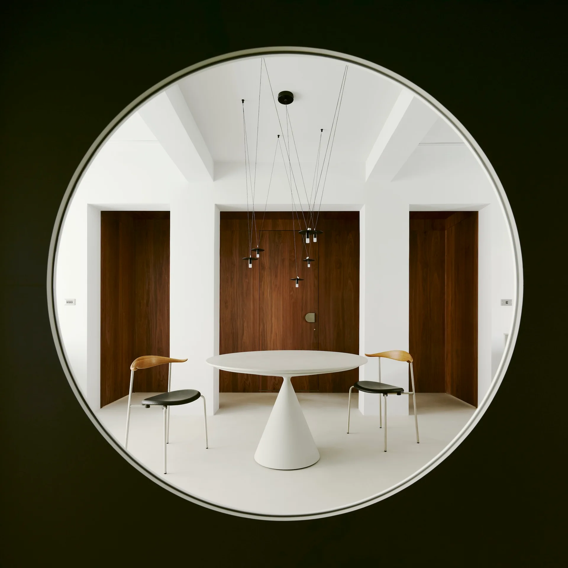 A minimalist dining room with a white pedestal table and two chairs is seen through a large circular cutout in a dark foreground wall.