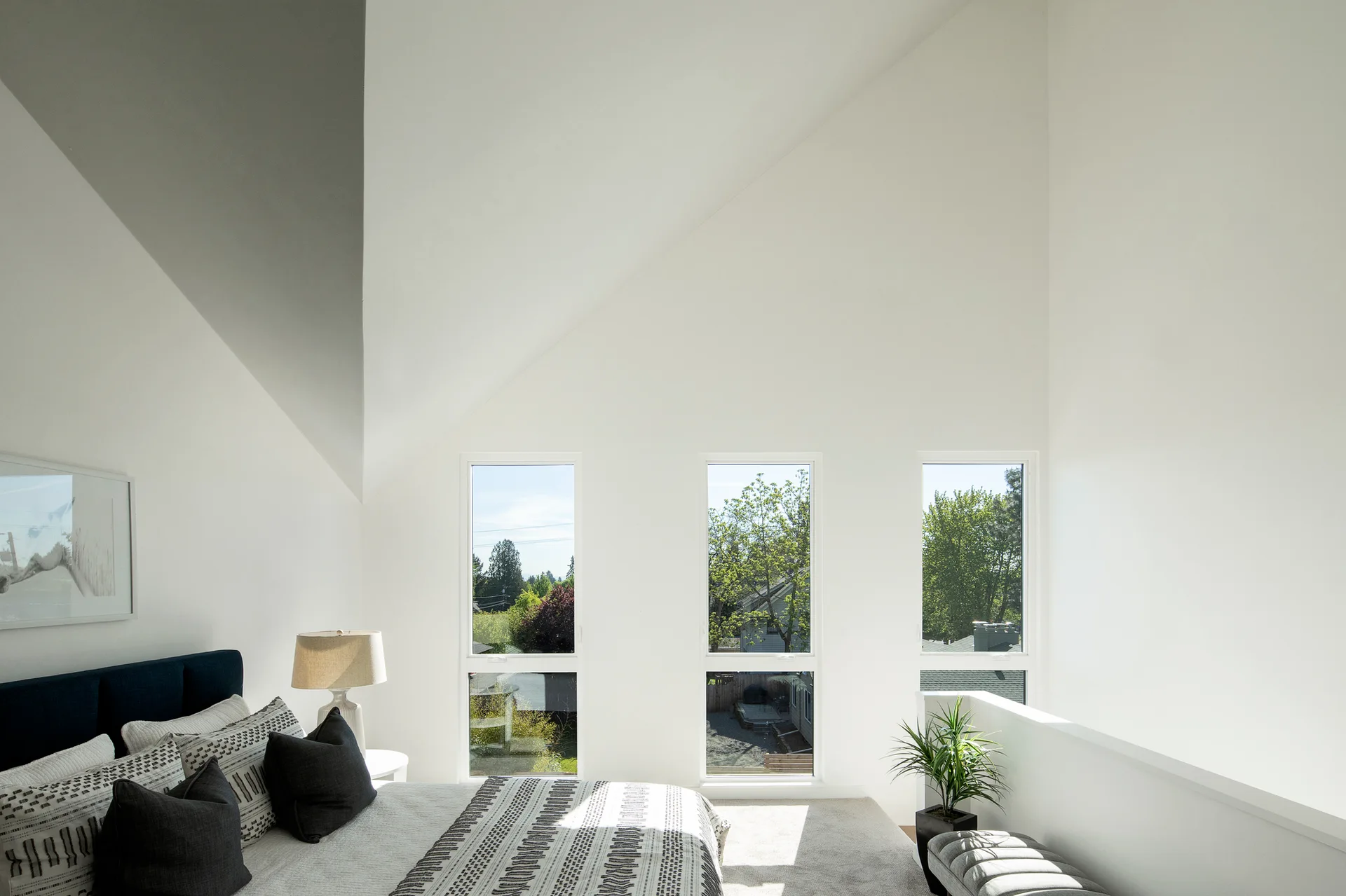 A bright bedroom featuring a vaulted white ceiling, three narrow vertical windows, a bed with patterned grey bedding, and a dark blue headboard.
