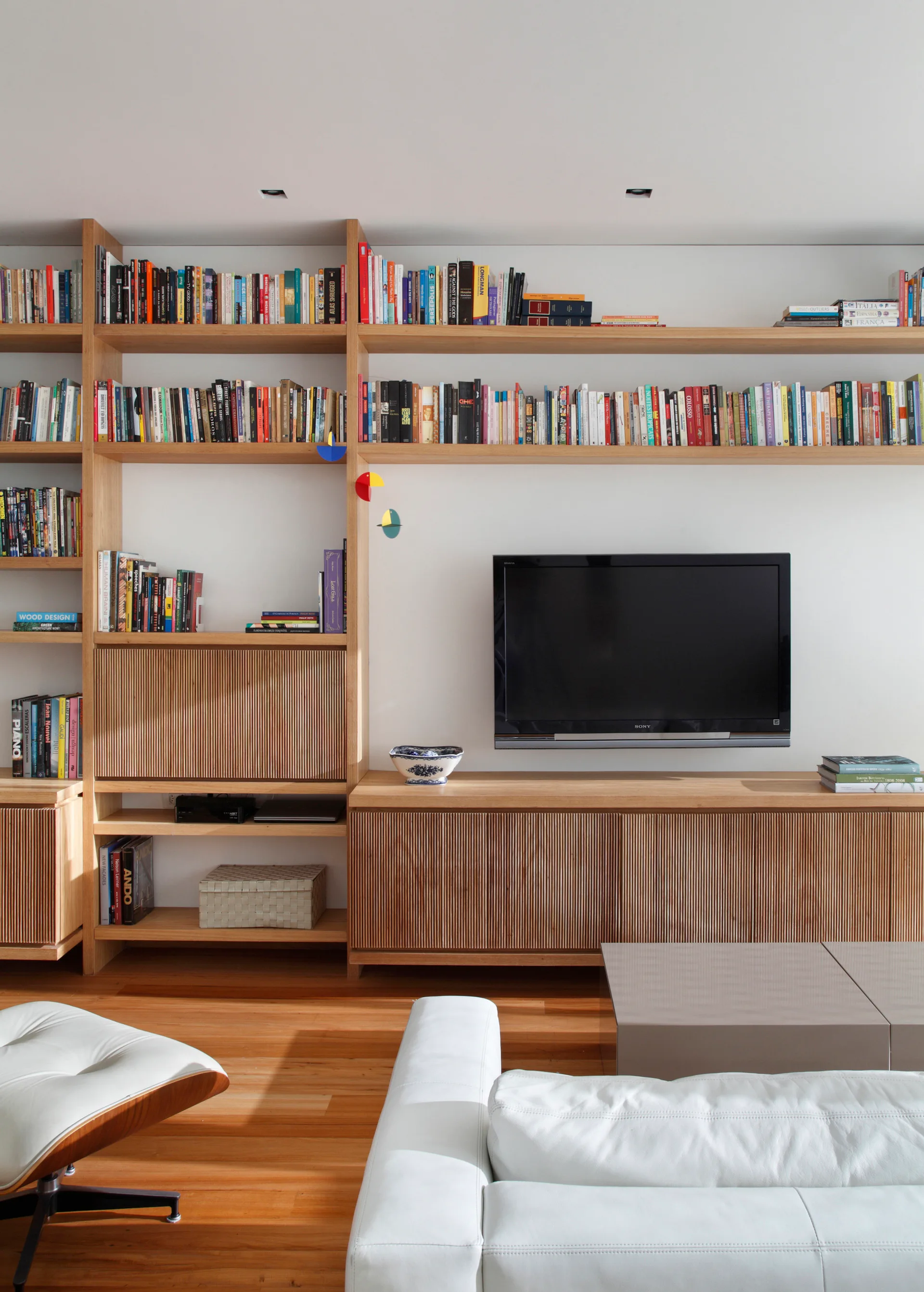 A large light wood bookshelf unit fills a wall with books, surrounding a wall-mounted TV and slatted wood cabinets above a white leather sofa.