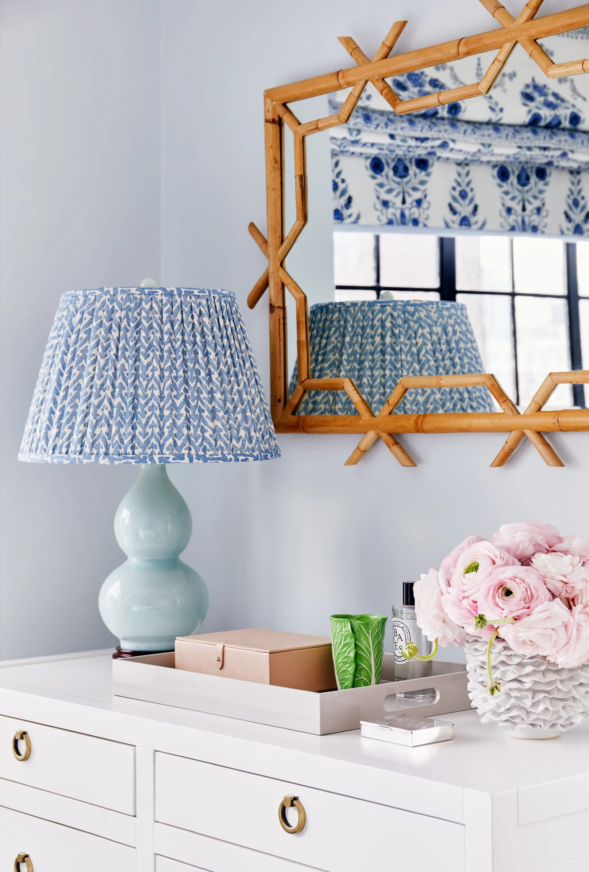 A white dresser styled with a blue ceramic lamp, a bamboo-framed mirror, and a tray of small decorative items next to pink flowers.