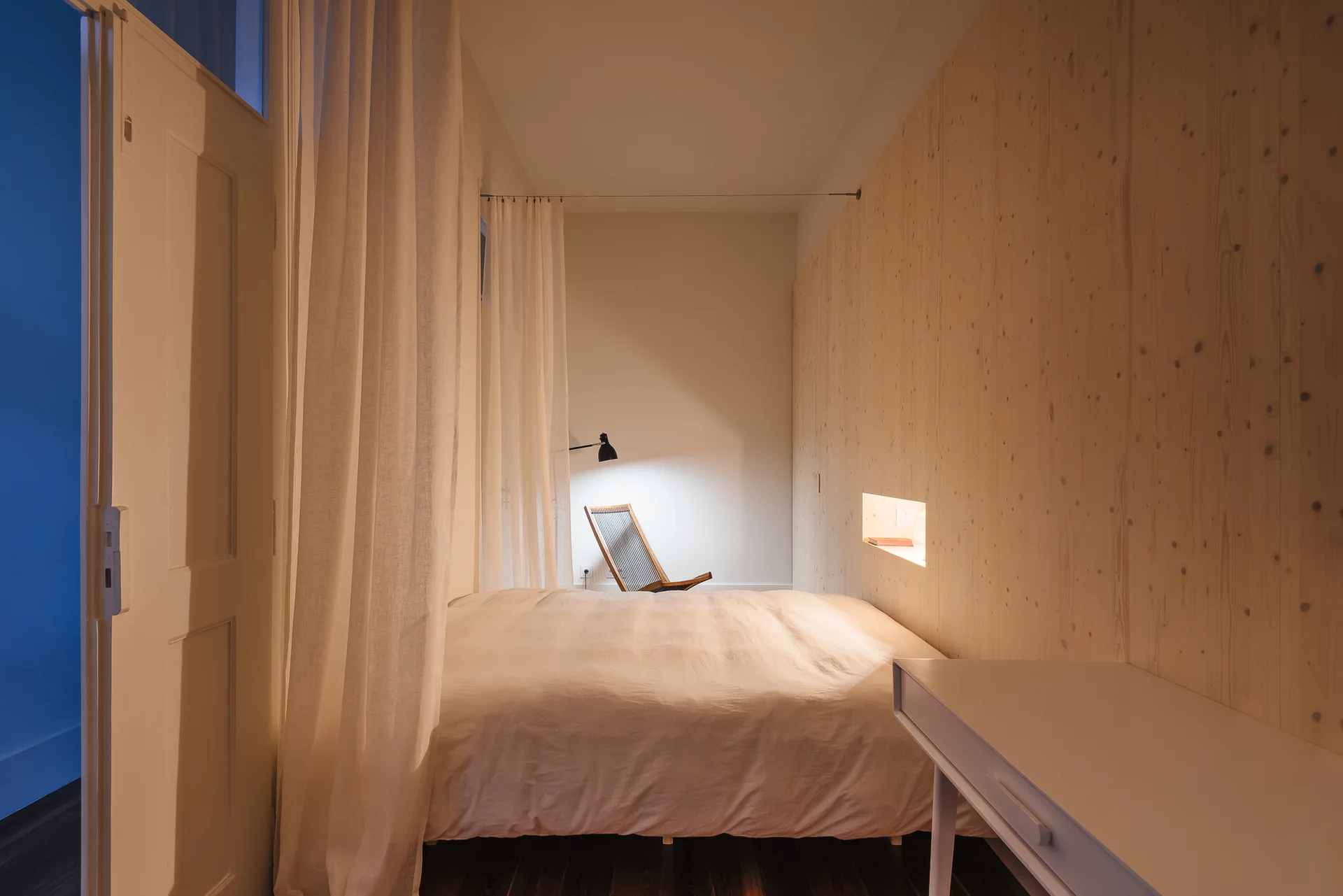 A minimalist bedroom featuring a bed with white linens next to a knotty pine paneled wall and a sheer white curtain partition.