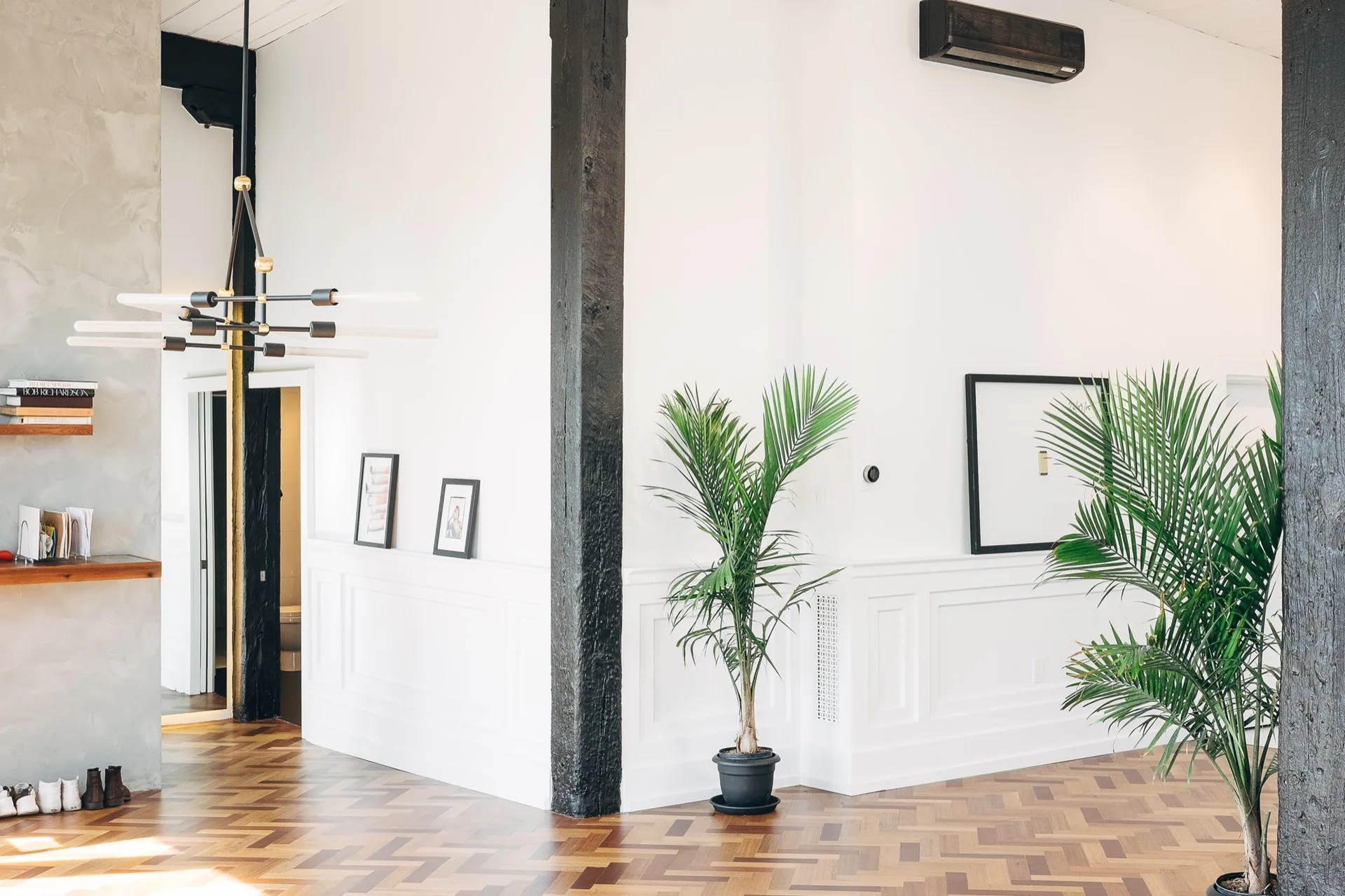 An interior space features white walls with wainscoting, dark timber columns, herringbone parquet flooring, and potted palm plants.